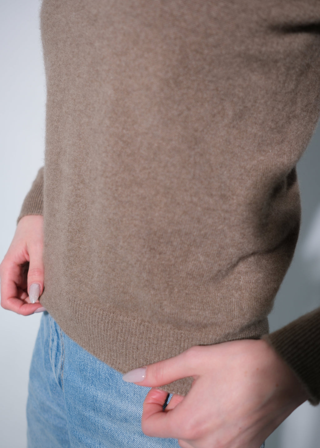 A close-up of a person wearing a taupe brown knit sweater and light blue jeans, showing the ribbed hem and hands adjusting the sweater.