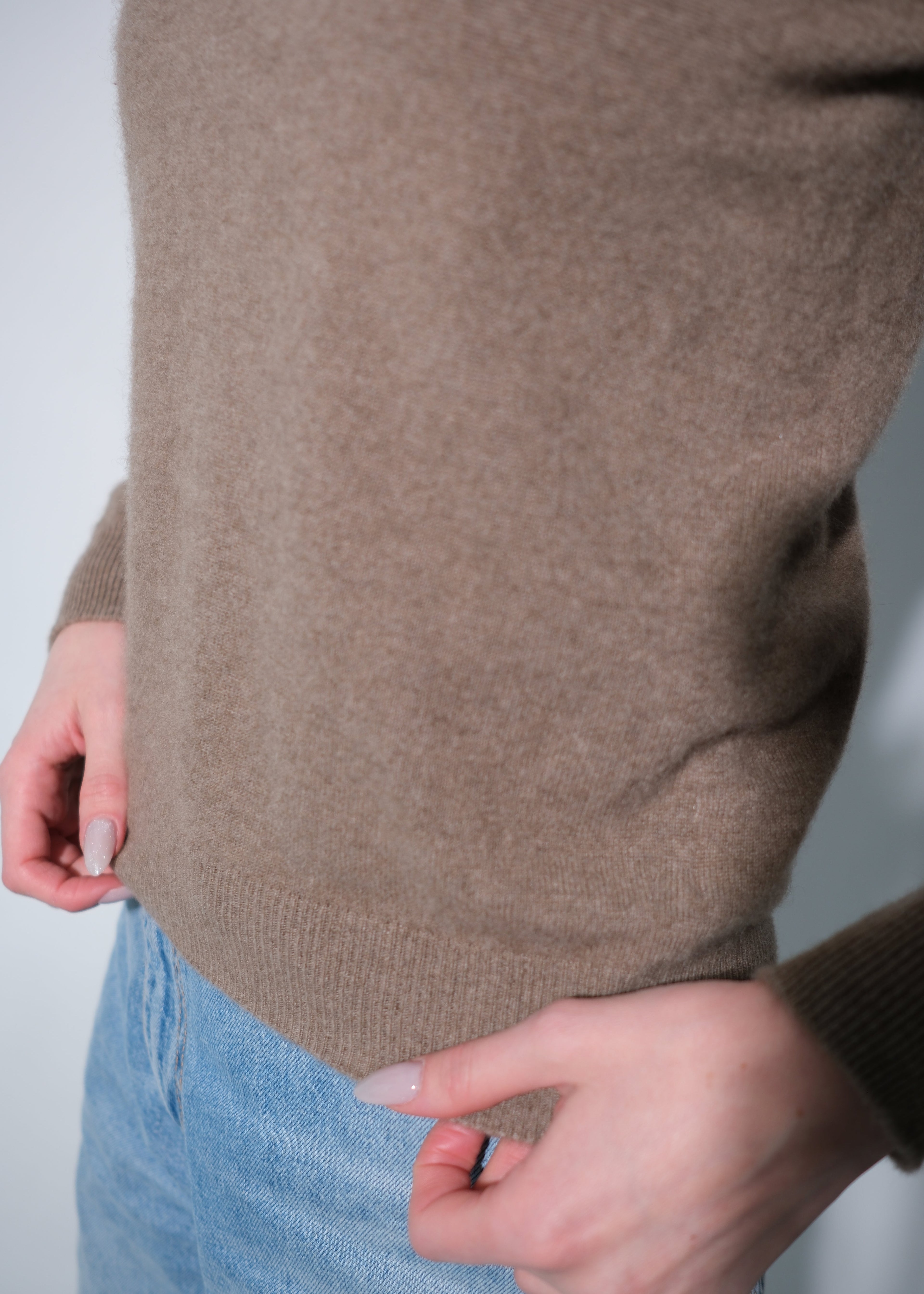 A close-up of a person wearing a taupe brown knit sweater and light blue jeans, showing the ribbed hem and hands adjusting the sweater.
