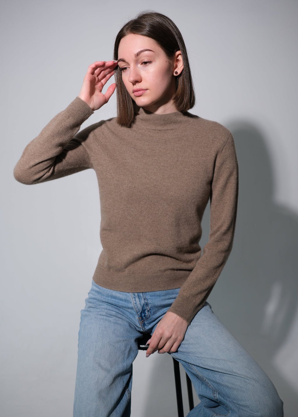 A woman wearing a brown mock-neck knit sweater and light blue jeans, posing while sitting on a stool against a plain gray background.