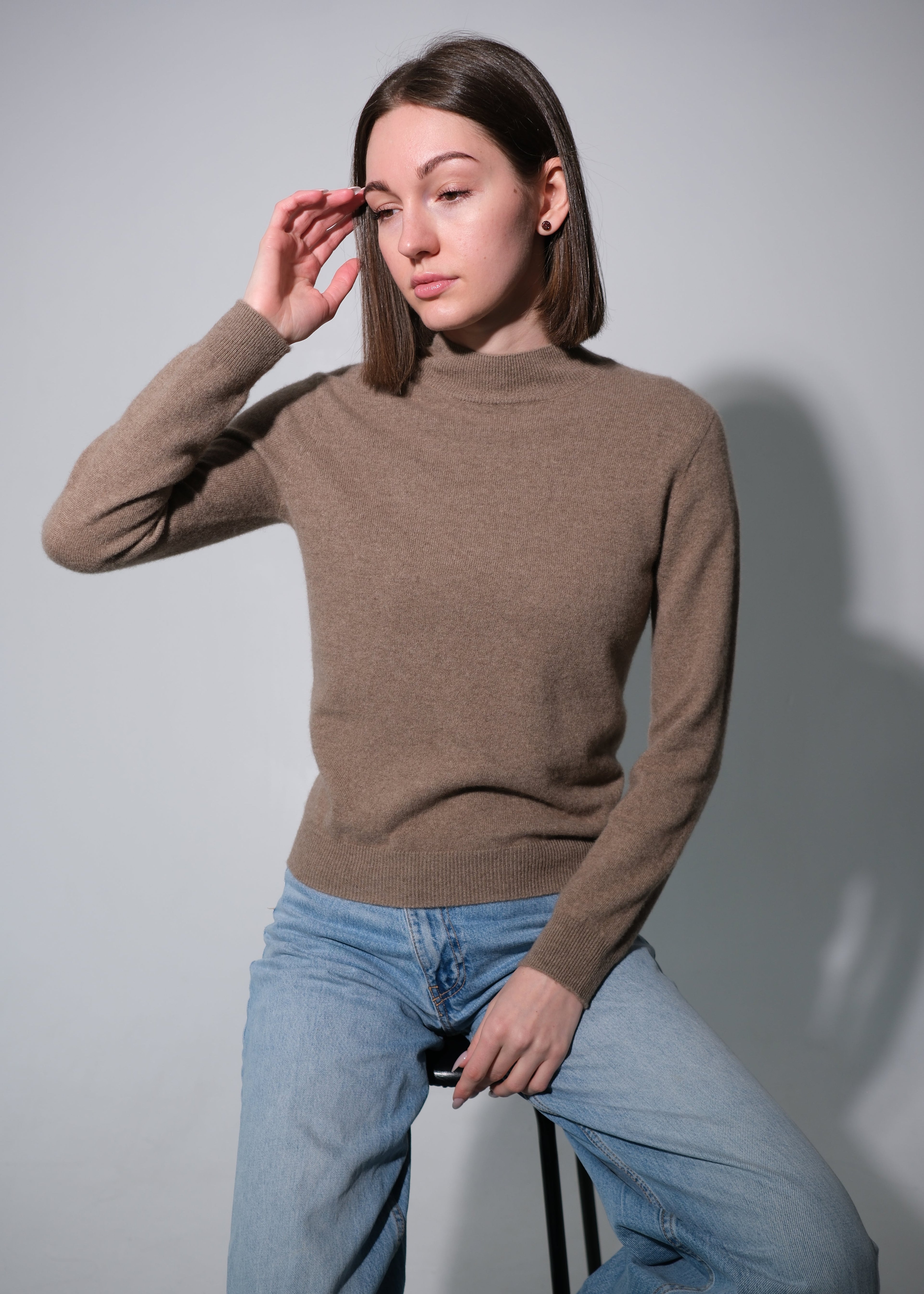 A woman wearing a brown mock-neck knit sweater and light blue jeans, posing while sitting on a stool against a plain gray background.