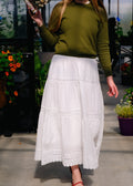 Person wearing a green sweater and white skirt outdoors with plants in the background