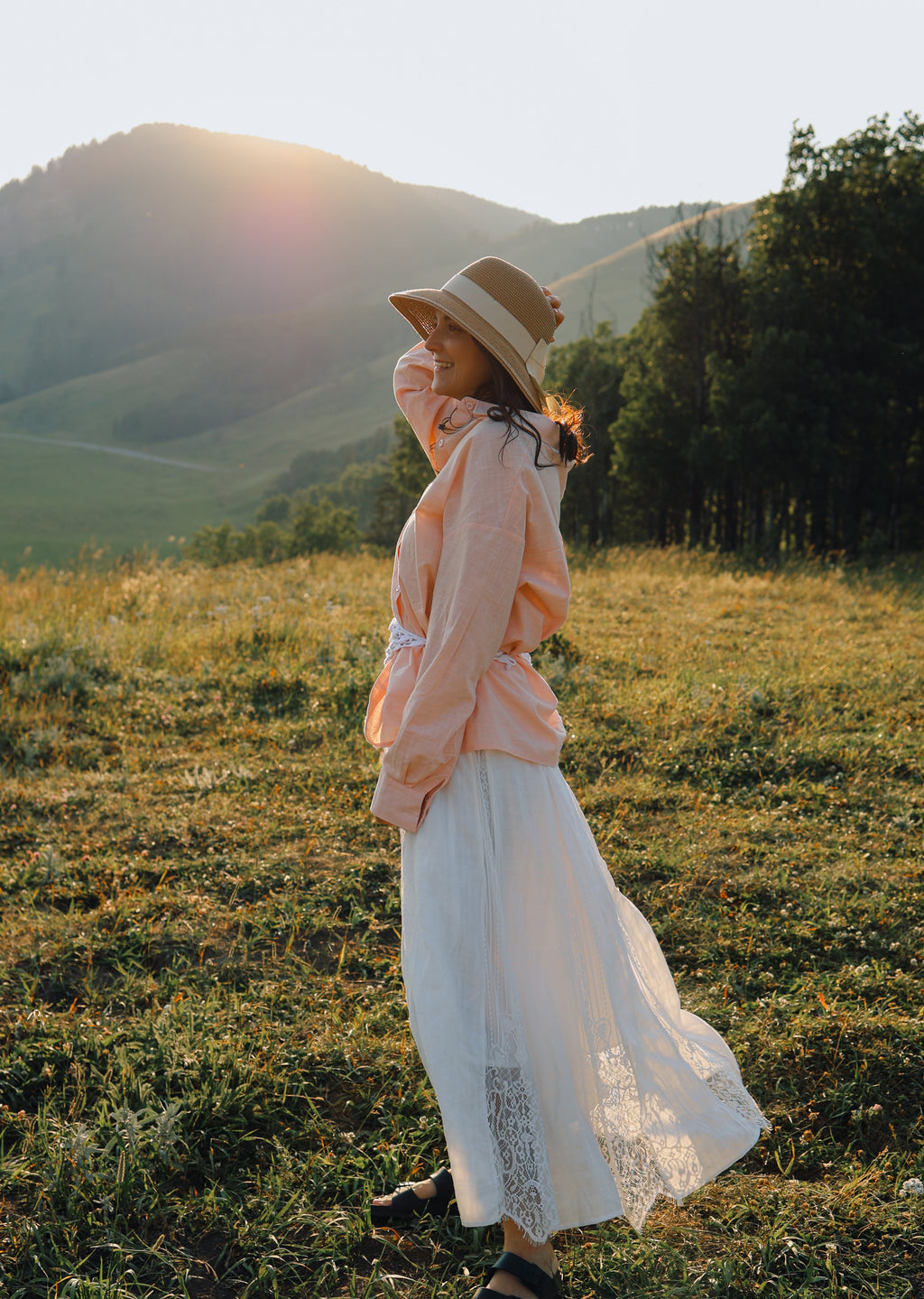 A woman stands in a grassy field at sunset wearing a wide-brim straw hat, a light peach long-sleeve top, and a long white skirt, with rolling hills and trees in the background.