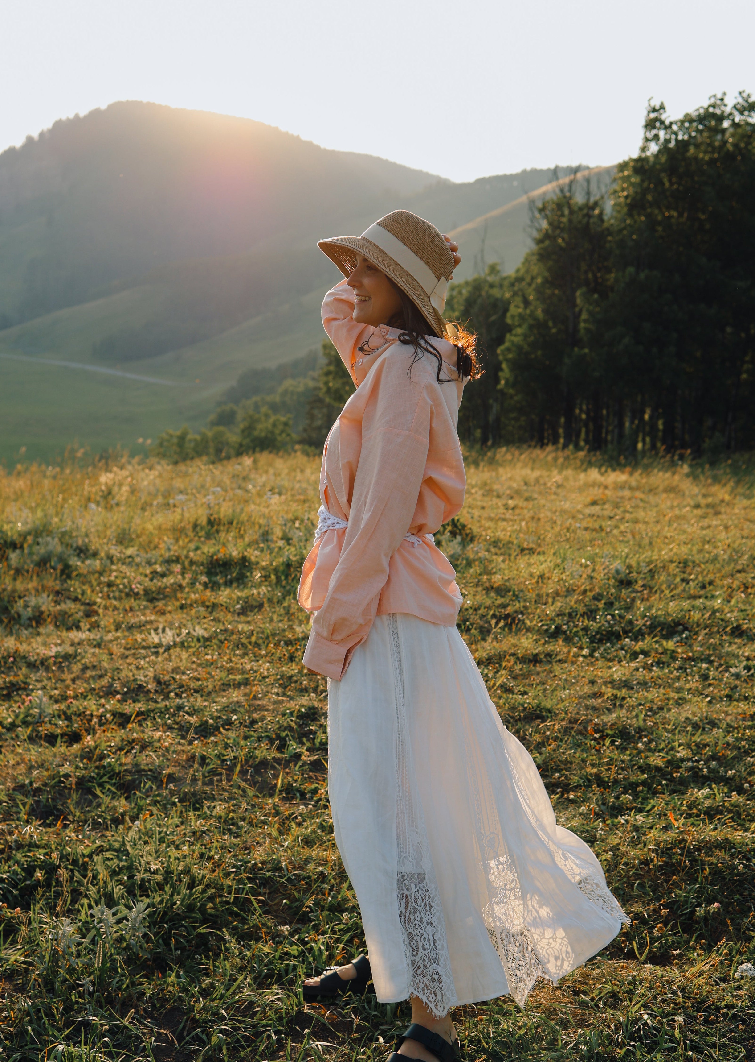 A woman stands in a grassy field at sunset wearing a wide-brim straw hat, a light peach long-sleeve top, and a long white skirt, with rolling hills and trees in the background.