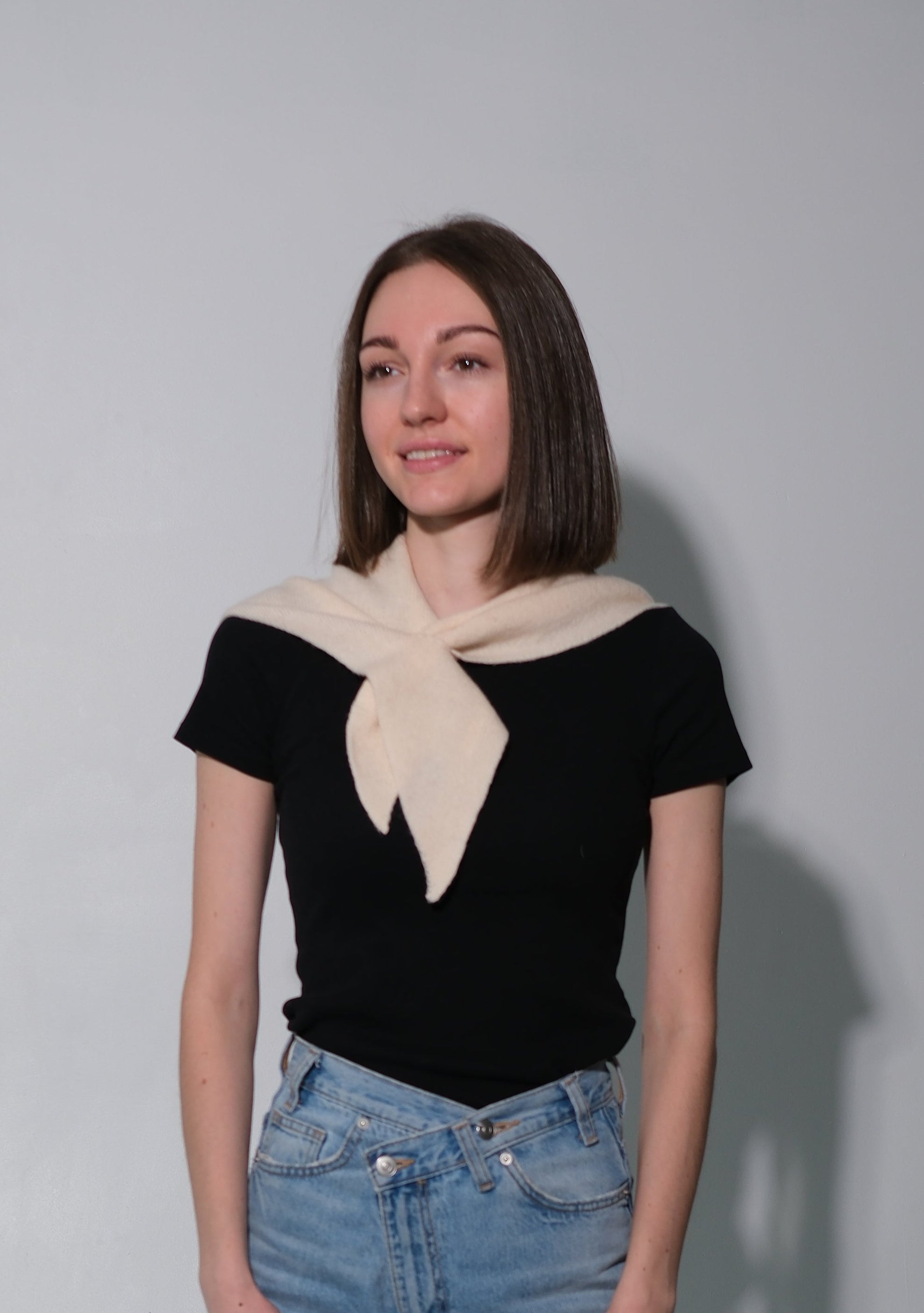 A woman stands against a plain light gray wall wearing a black short-sleeve top with a cream-colored scarf tied around the shoulders, light-wash wide-leg jeans, and gray socks.