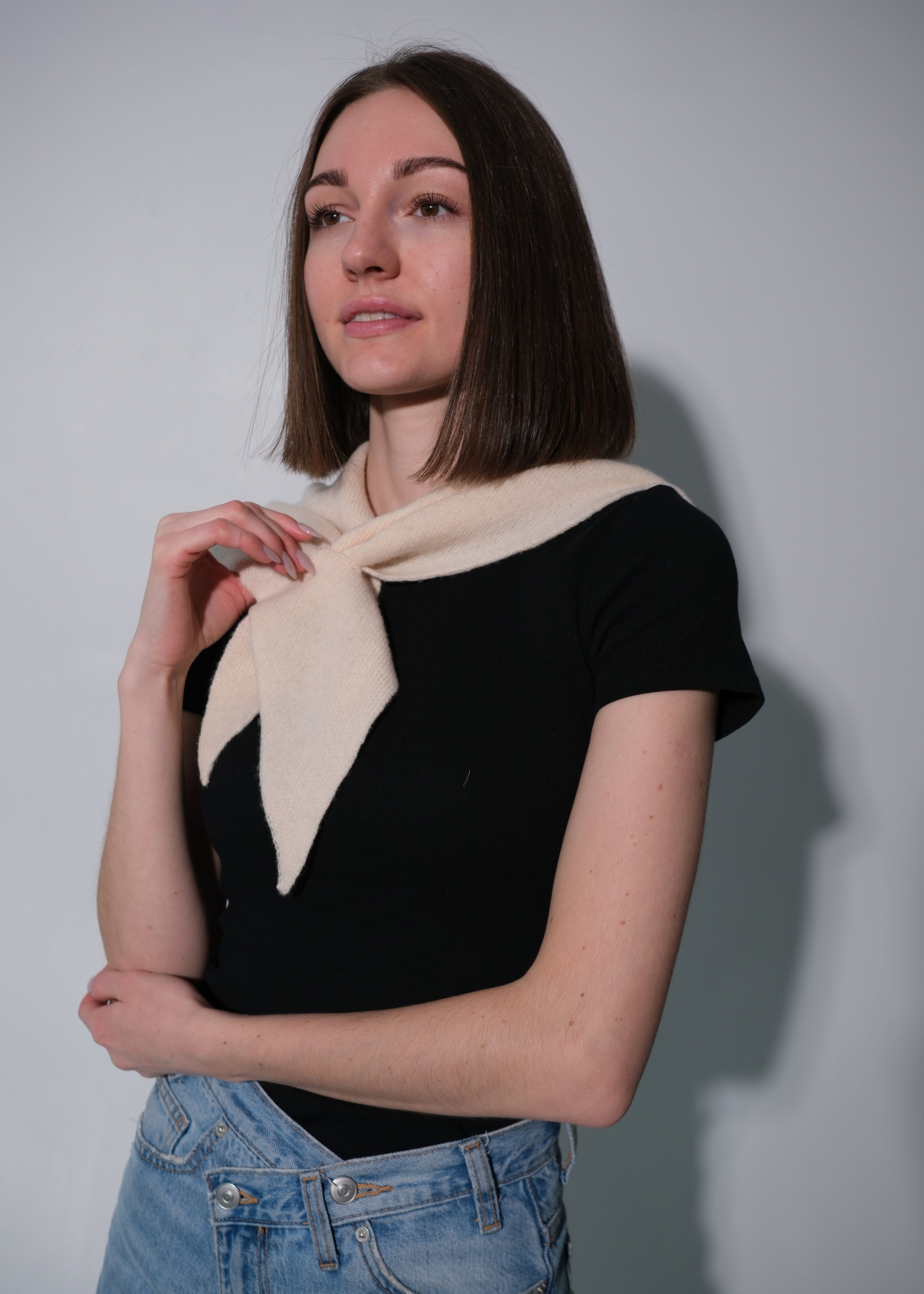 A woman with shoulder-length brown hair wearing a black short-sleeve top and light blue jeans, modeling a cream-colored knit scarf tied around her neck against a plain gray studio background.
