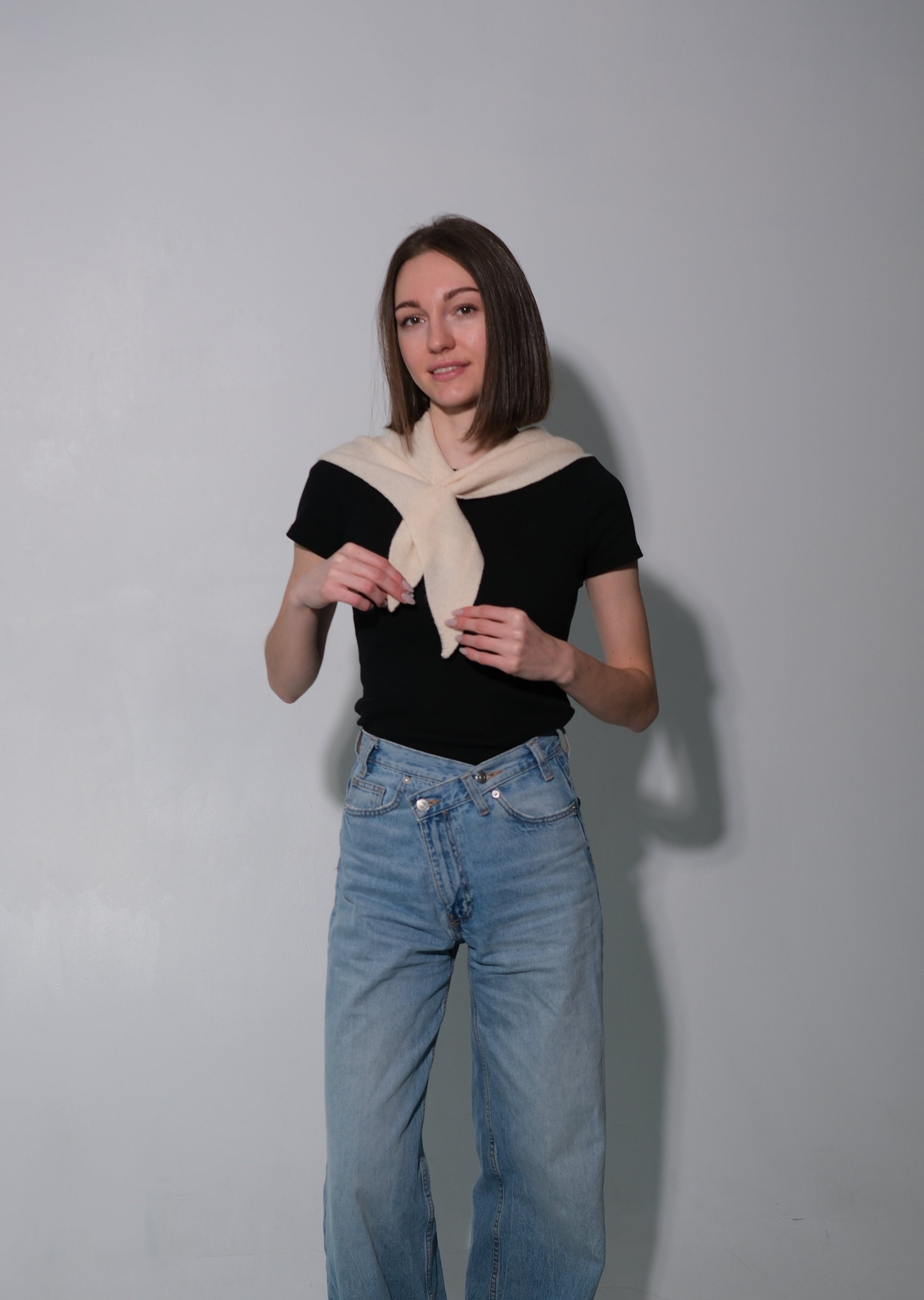 A woman modeling a black short-sleeve top with a cream knit collar, paired with light-wash wide-leg jeans, standing against a plain gray studio background.