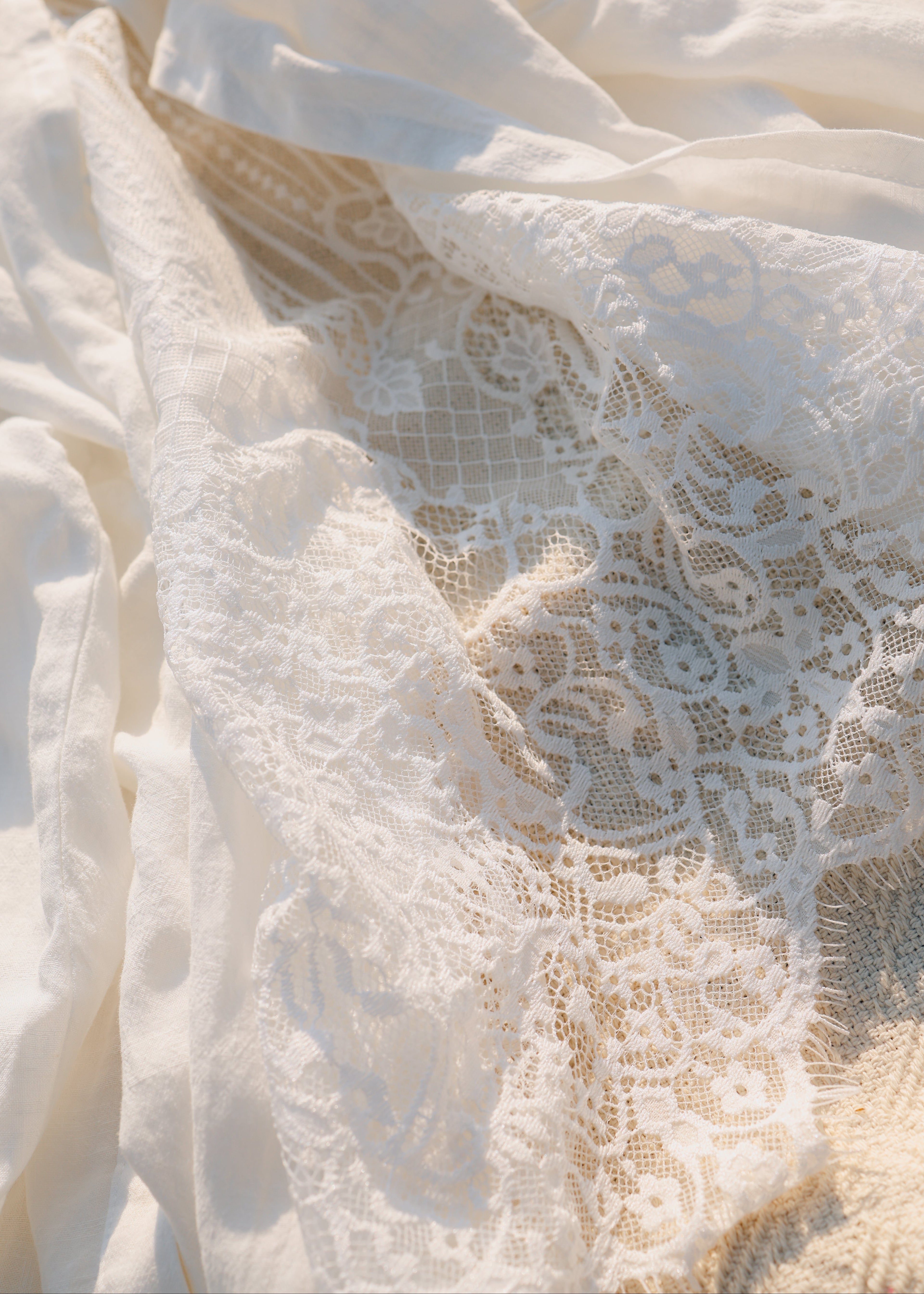 Close-up of white lace fabric draped over a soft beige textile, showing detailed floral lace pattern and light filtering through the folds.