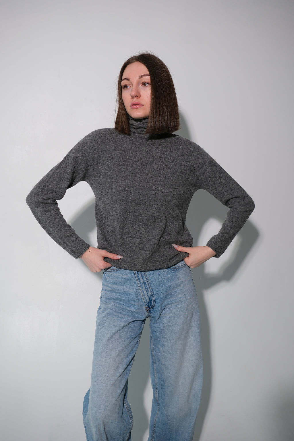 A woman with shoulder-length brown hair wearing a dark gray turtleneck sweater and light blue jeans, posing with her hands on her hips against a plain light gray wall.