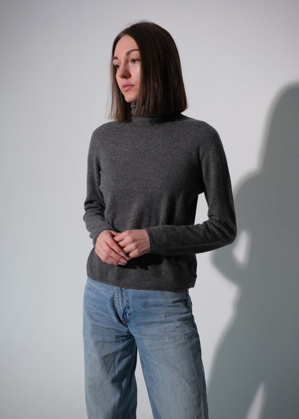A woman wearing a dark gray mock neck knit sweater and light blue jeans, standing against a light gray wall with a soft shadow cast on the right.