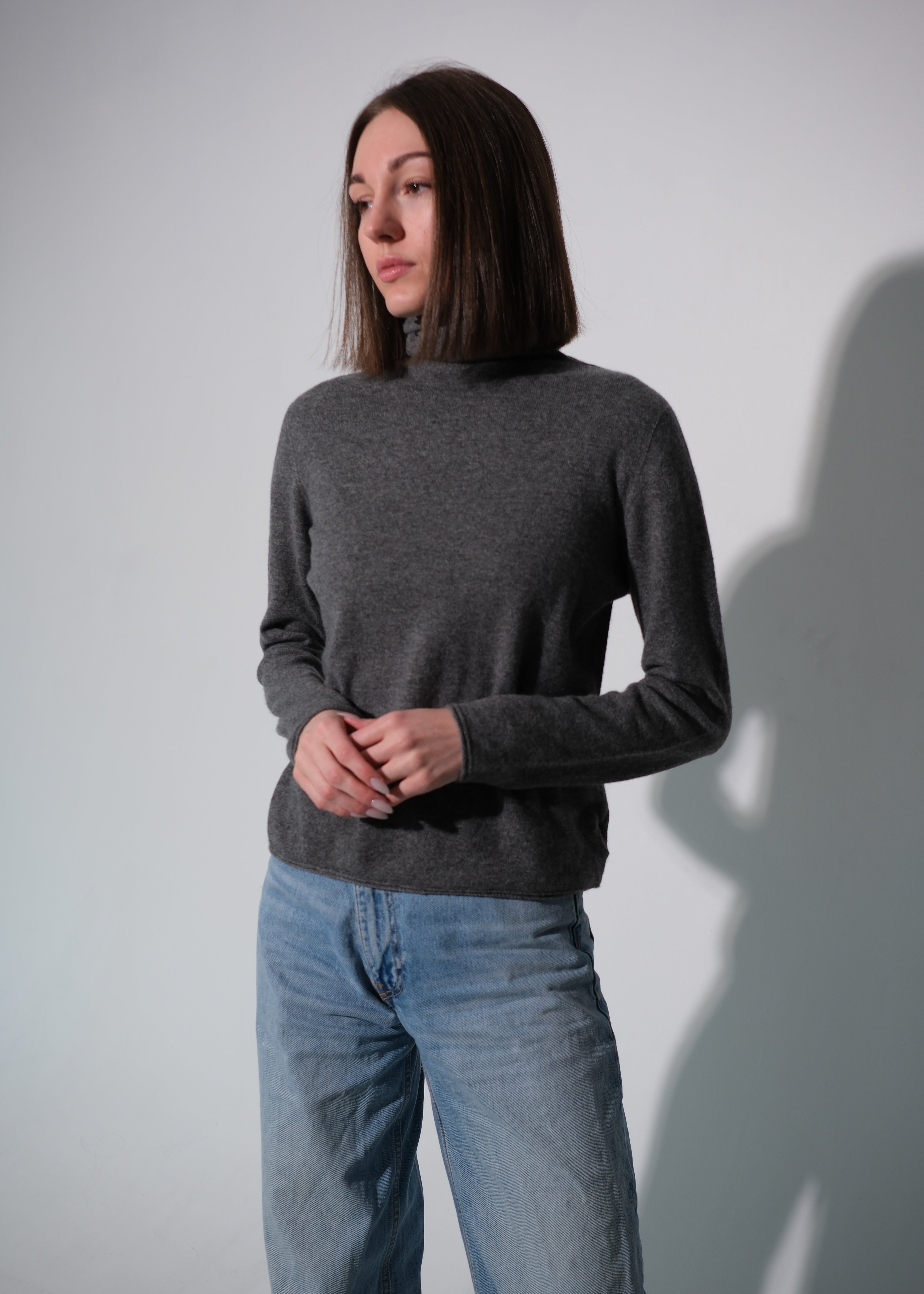 A woman wearing a dark gray mock neck knit sweater and light blue jeans, standing against a light gray wall with a soft shadow cast on the right.