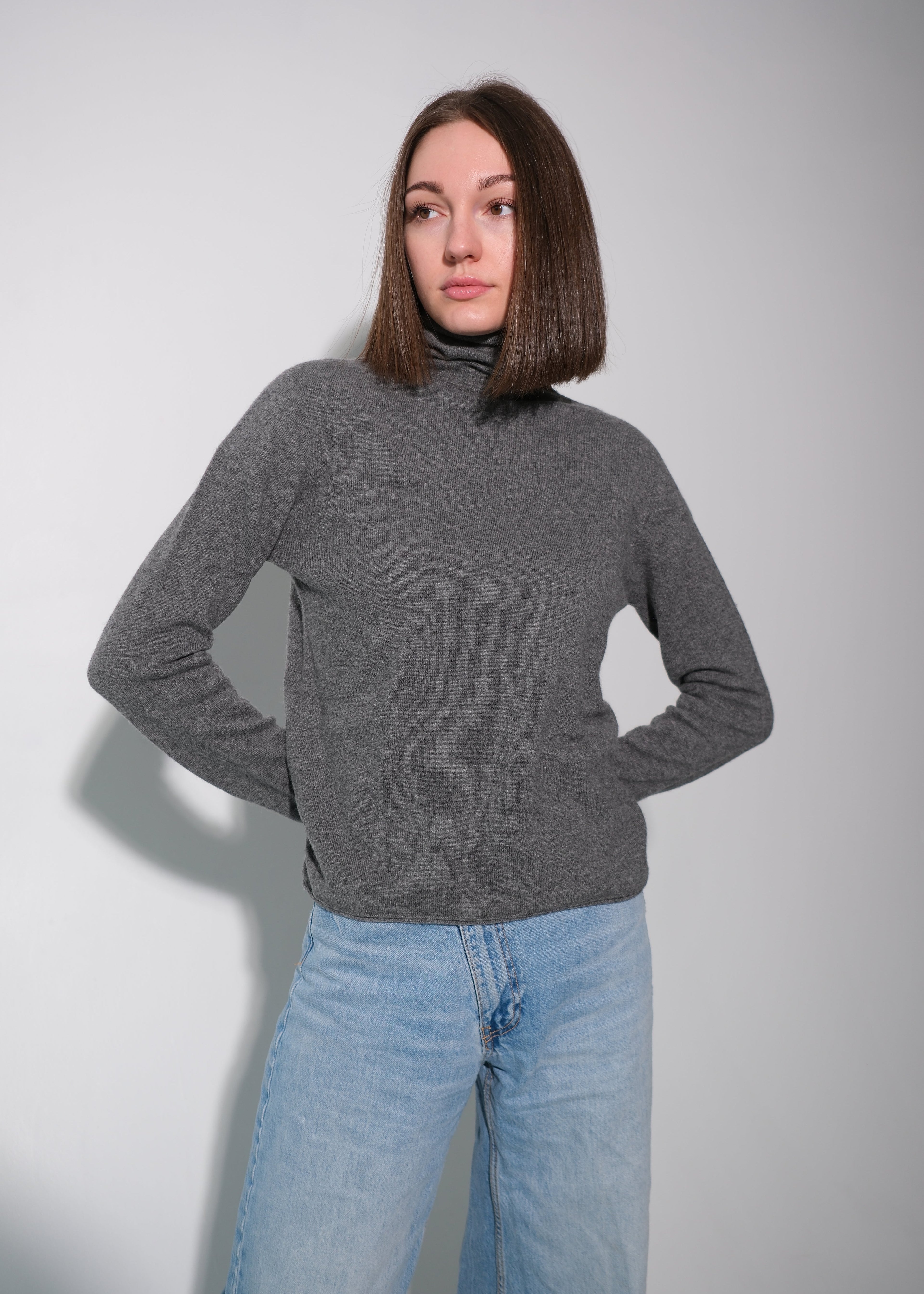 A woman with shoulder-length brown hair wearing a dark gray ribbed turtleneck sweater and light blue wide-leg jeans, standing against a light gray studio background.