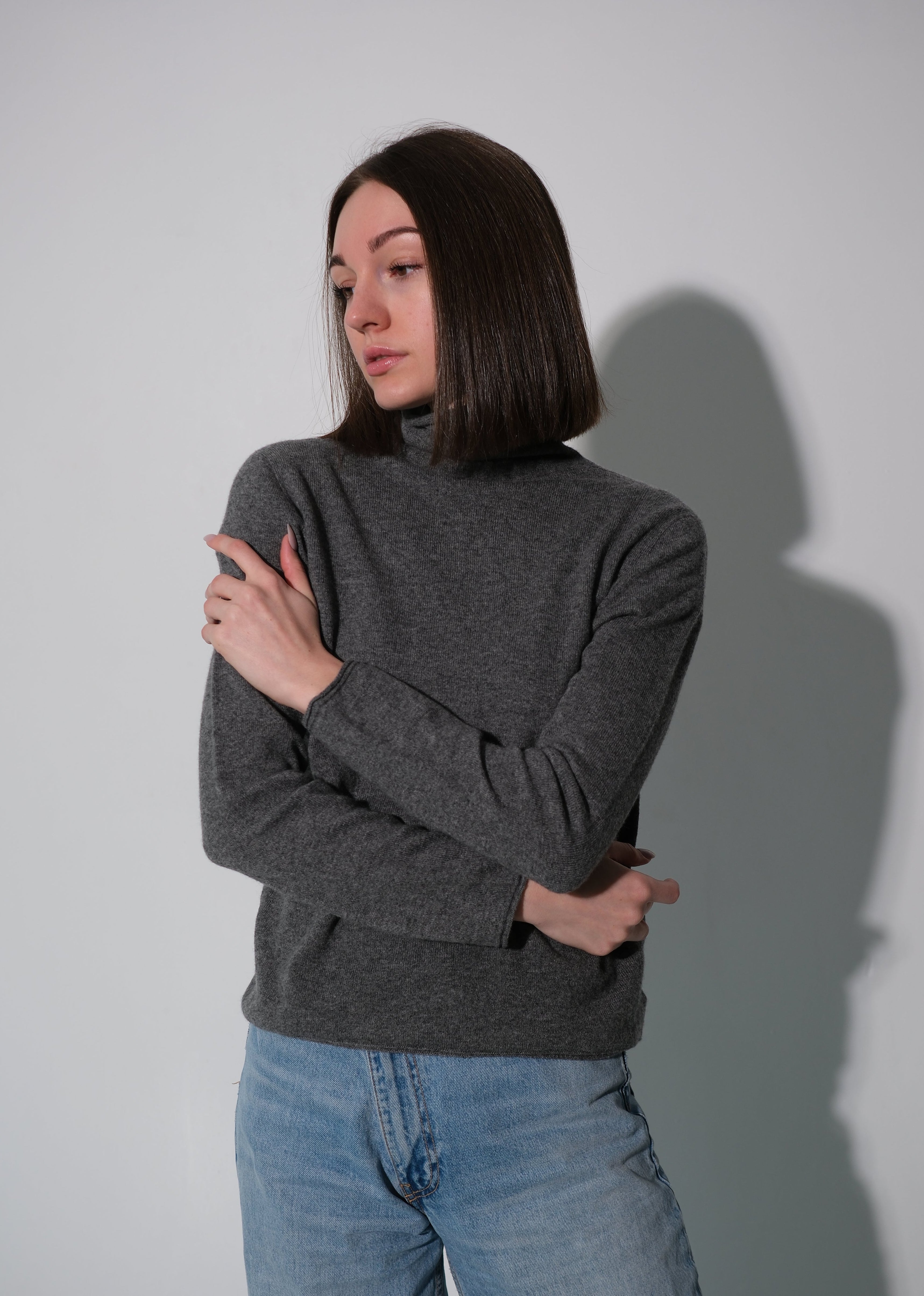 A woman with dark shoulder-length hair wearing a dark gray mock neck knit sweater and light blue jeans, standing against a light gray wall with a soft shadow.