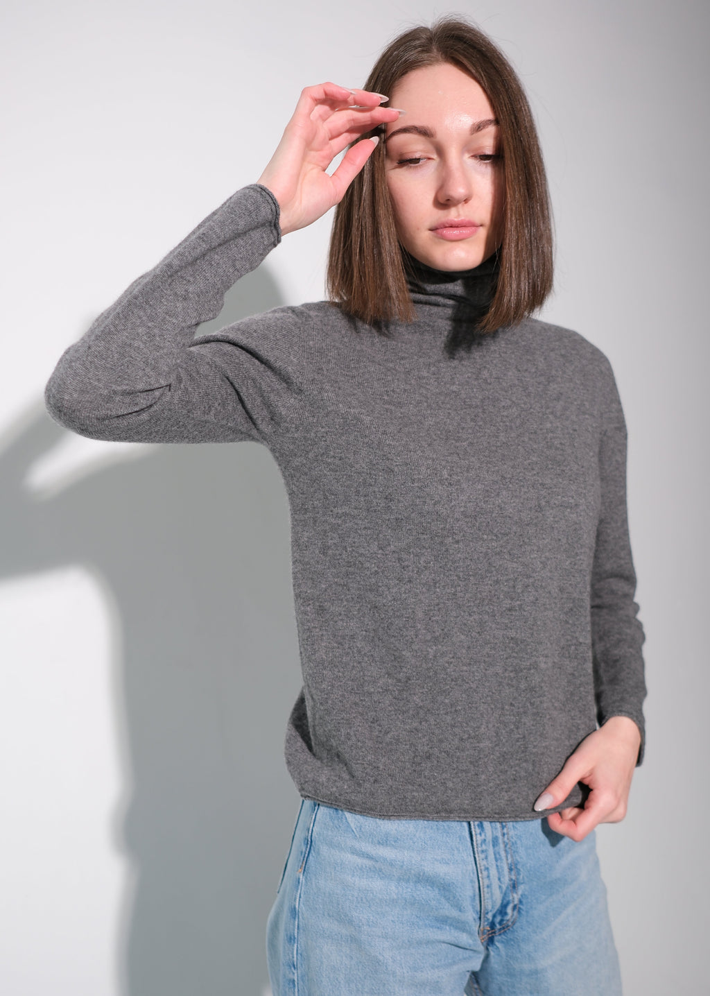 A woman wearing a gray long-sleeve turtleneck sweater and light blue jeans, posing against a light gray studio background.