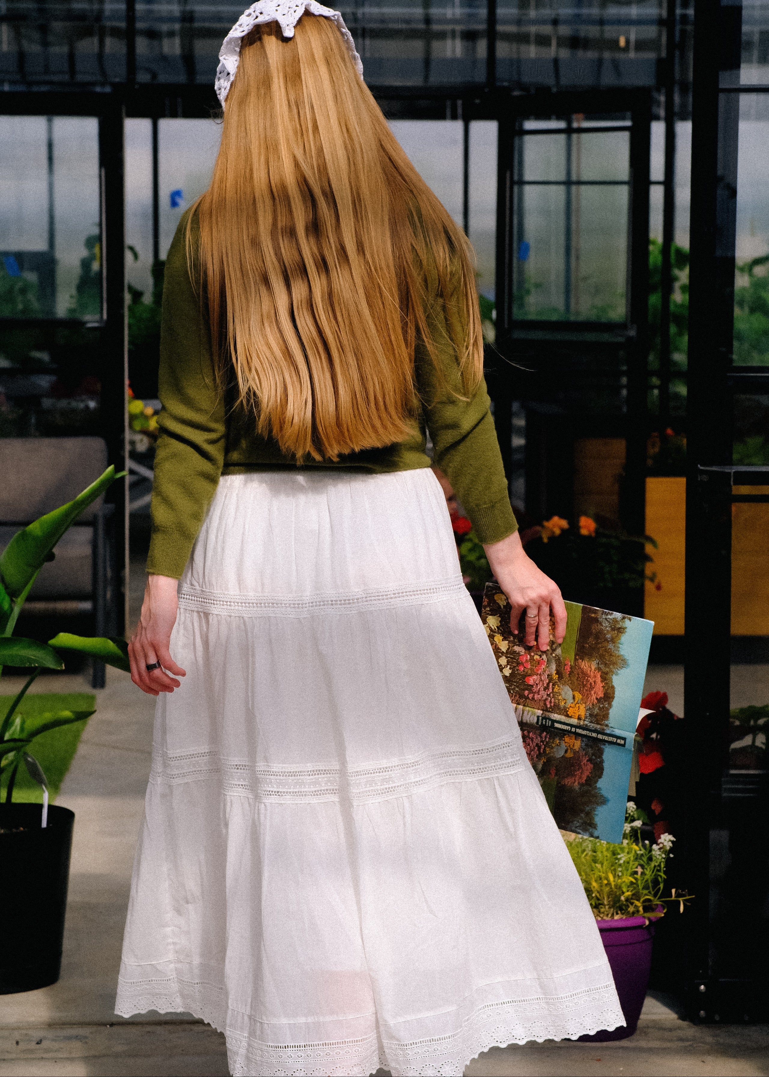 Person with long blonde hair wearing a green top and white skirt, standing in an indoor setting.