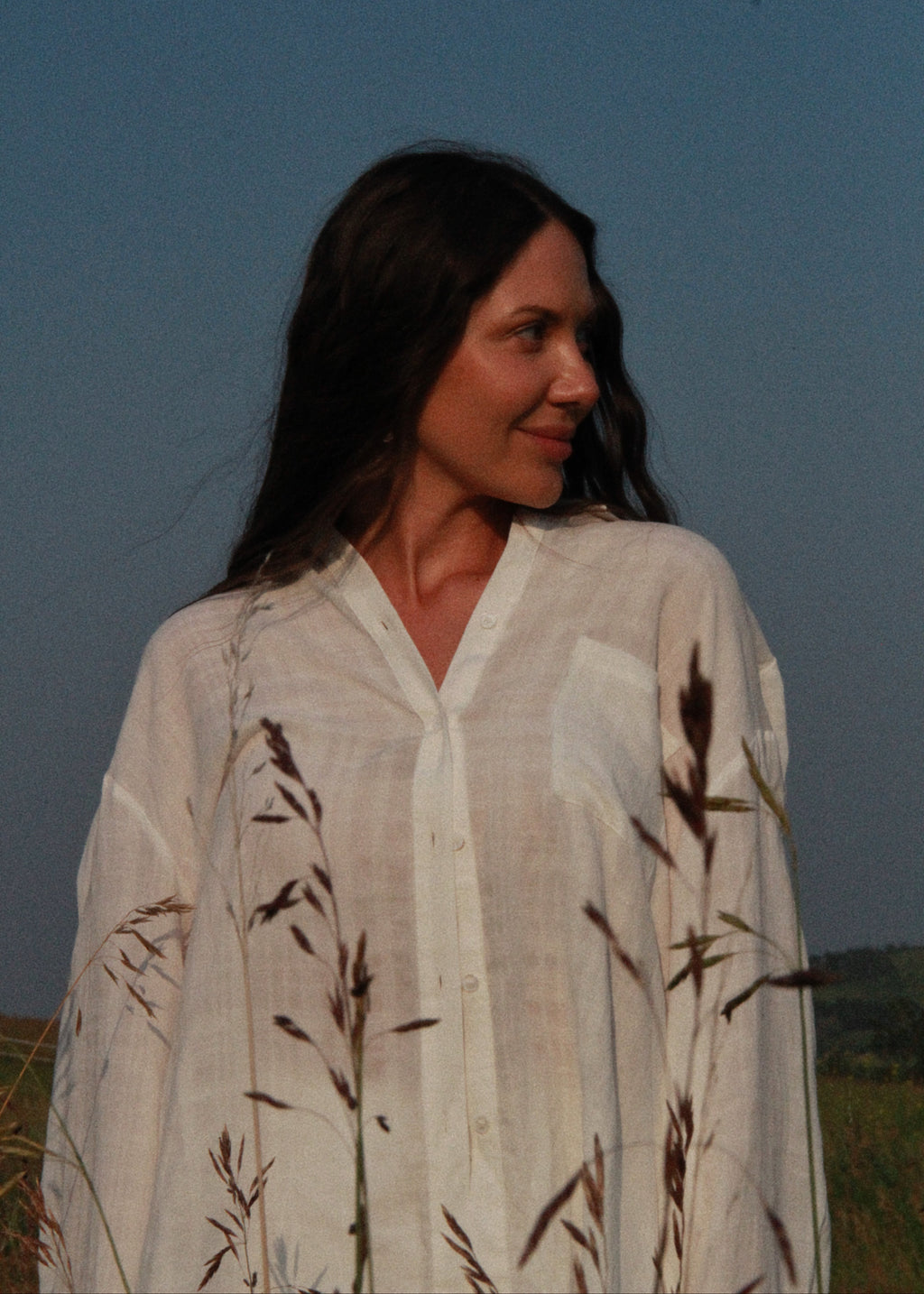 A woman wearing a loose white linen button-up shirt with dark brown grass motifs, standing outdoors against a blue sky.