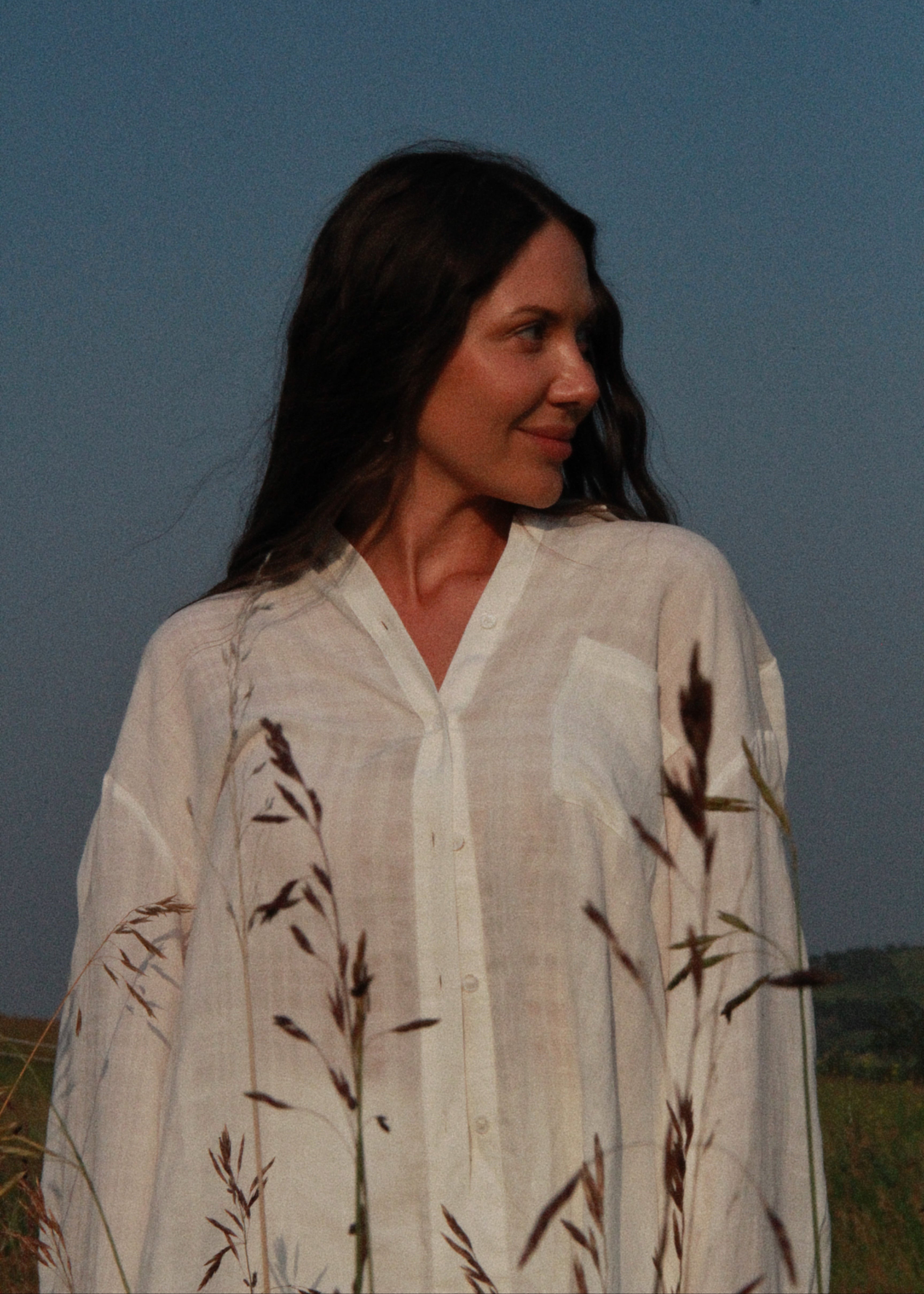 A woman wearing a loose white linen button-up shirt with dark brown grass motifs, standing outdoors against a blue sky.
