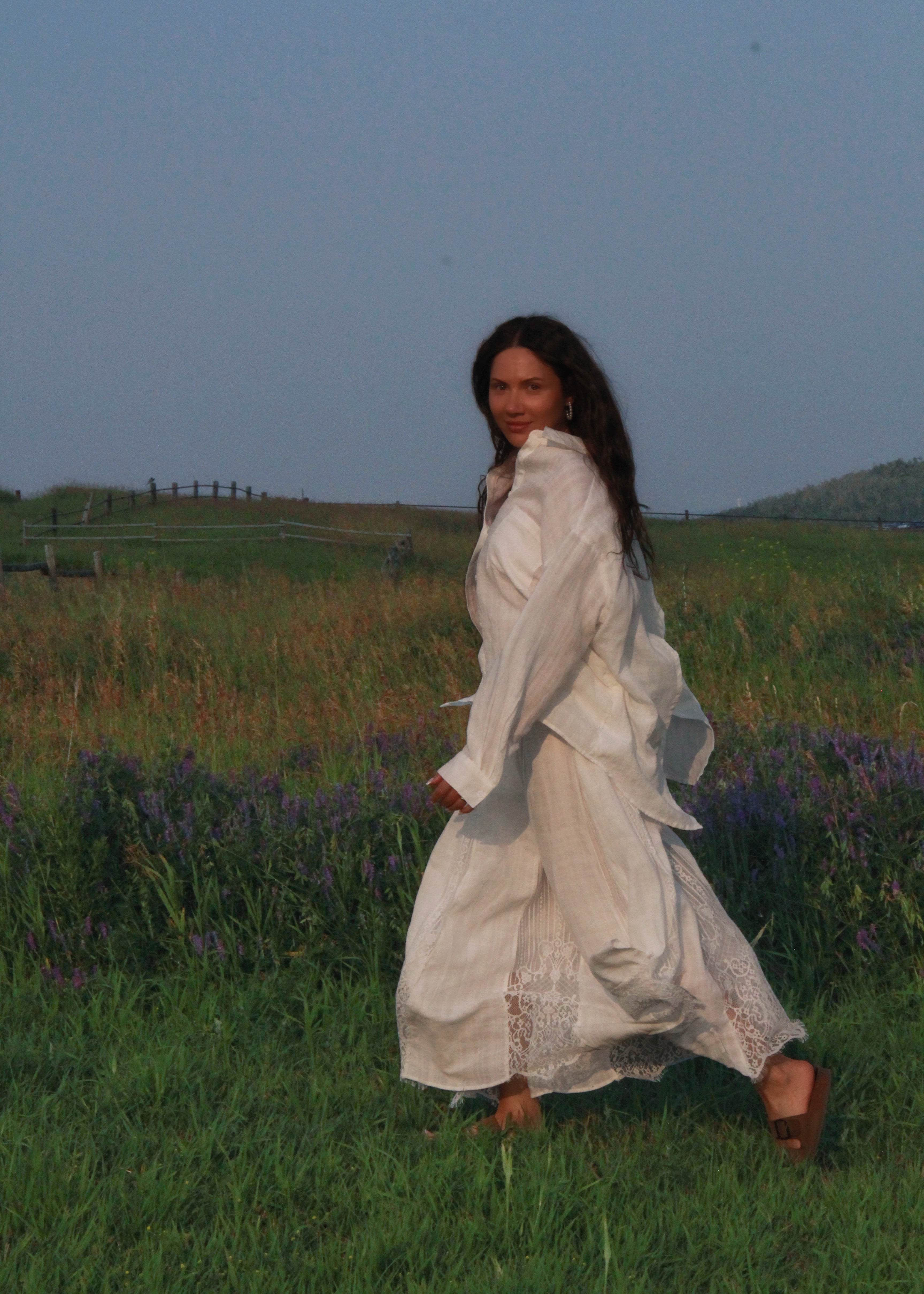 A woman wearing a long white dress stands in a grassy field with purple flowers under a blue sky.