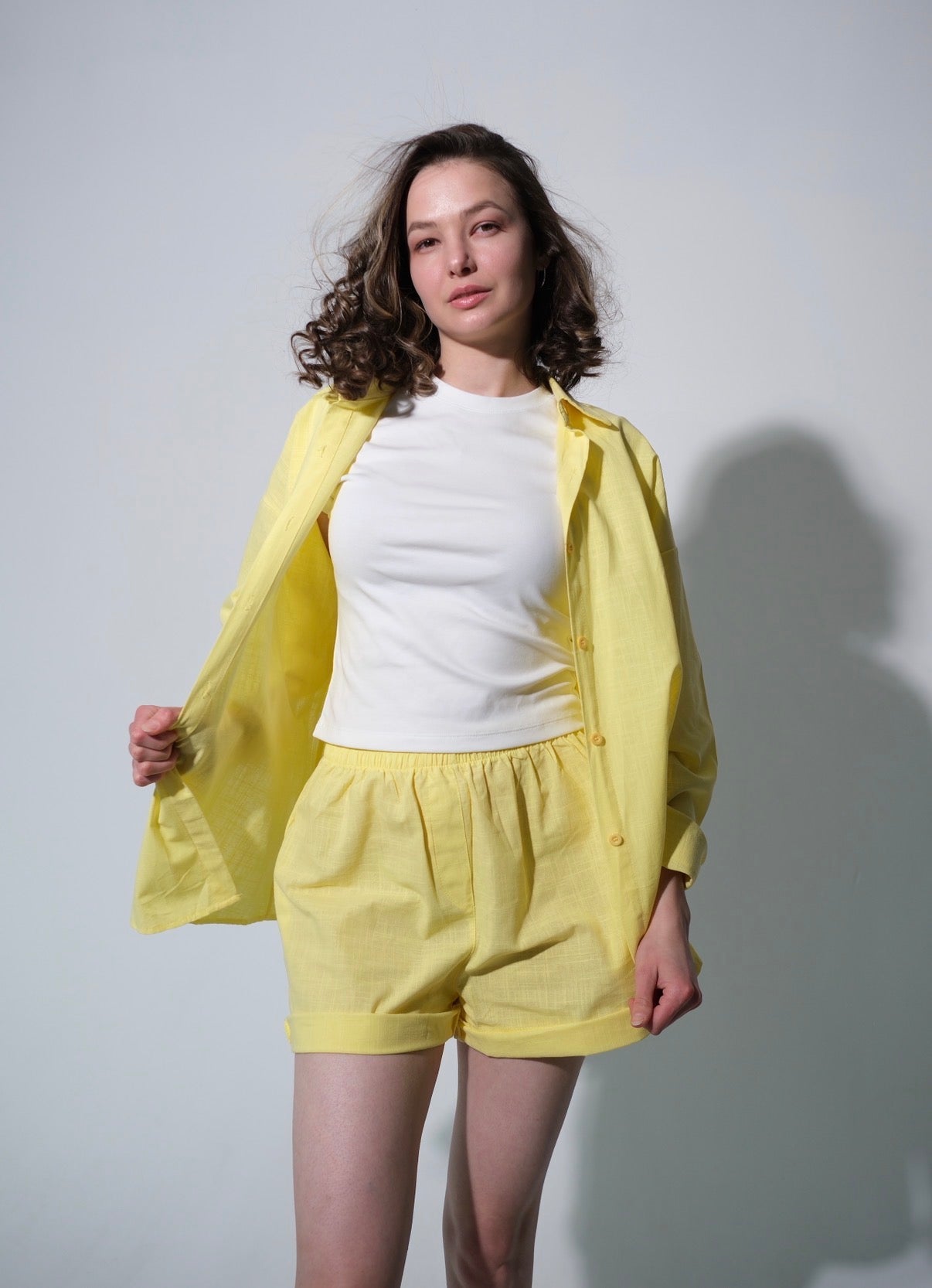 Woman wearing a relaxed yellow cotton matching set featuring an oversized button-up shirt and comfortable shorts. Lightweight summer outfit styled with a white top, photographed in a minimalist studio setting with soft natural lighting. Casual resort wear perfect for warm-weather days, vacations, and everyday comfort.