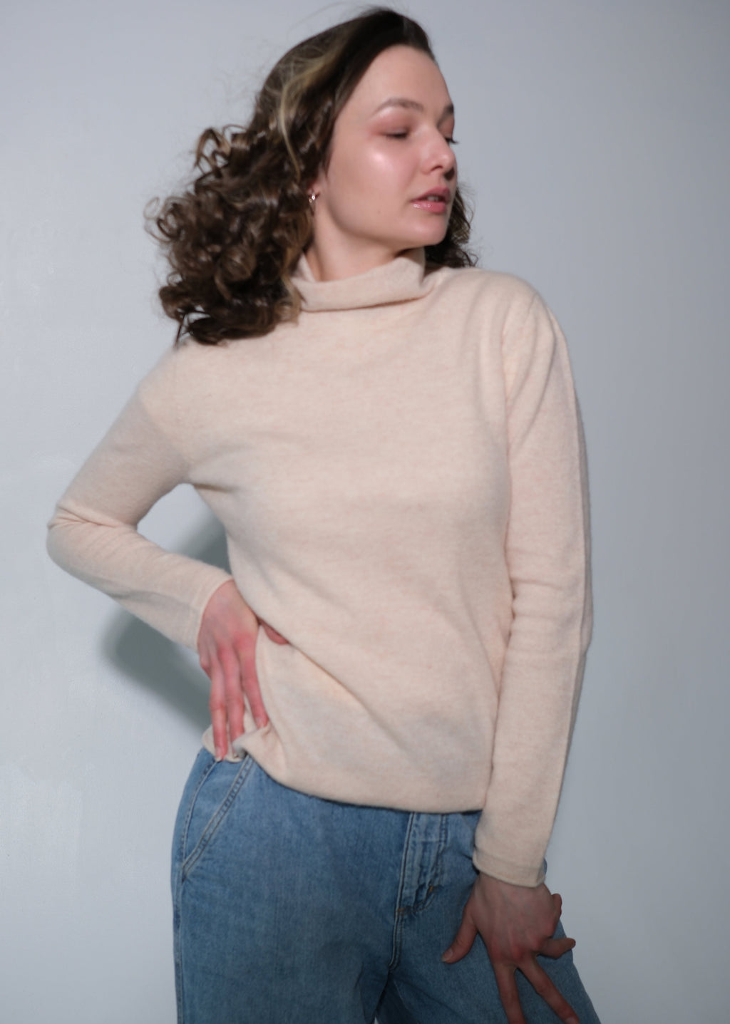 A woman wearing a light beige turtleneck sweater and blue jeans, posing against a plain light gray studio background.