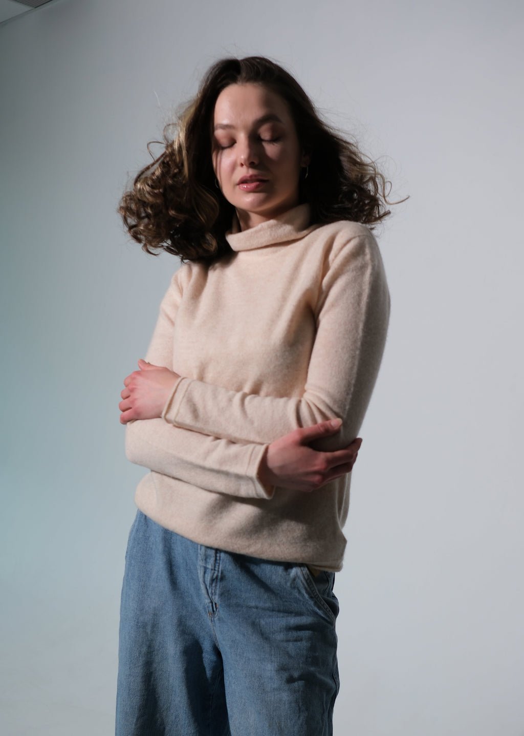 A woman with wavy brown hair stands with arms crossed, wearing a light beige turtleneck sweater and blue jeans against a plain light gray studio background.