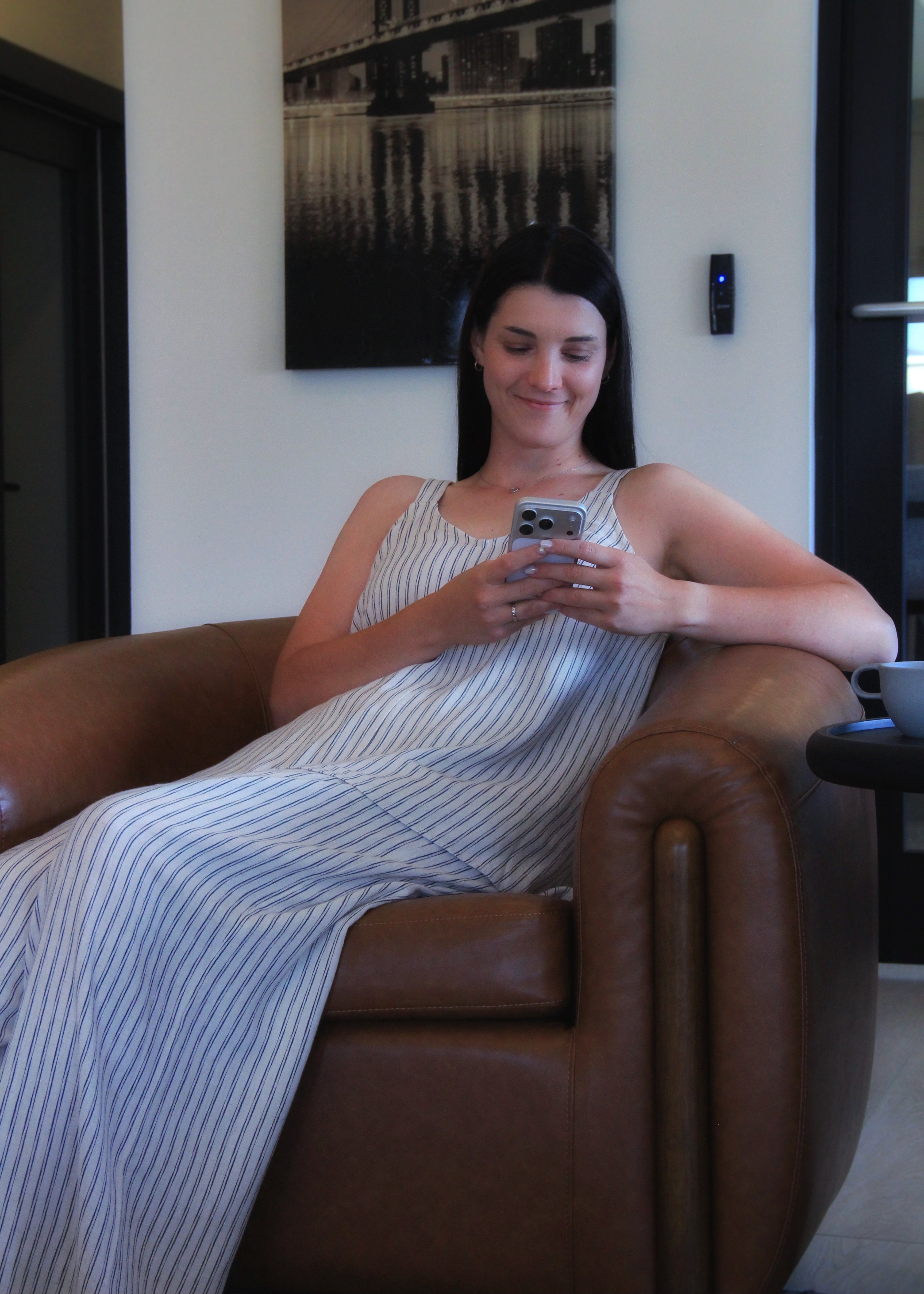 A woman with dark hair sits in a brown leather armchair, wearing a white sleeveless dress with thin blue vertical stripes and looking at her phone. A black and white bridge print hangs on the wall behind her.