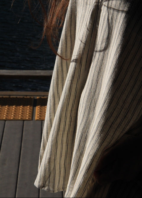 A close-up of a cream-colored linen fabric with thin vertical dark stripes, draped in soft folds with sunlight and shadows, set against a dark background.