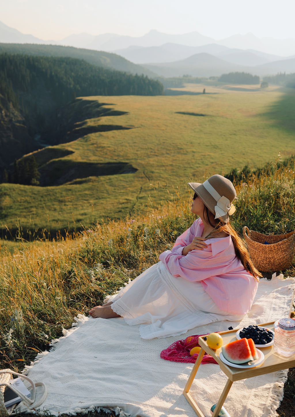 A woman in a pink kimono and straw hat sits on a white blanket in a mountain meadow, with a small picnic table holding fruit and a basket nearby.