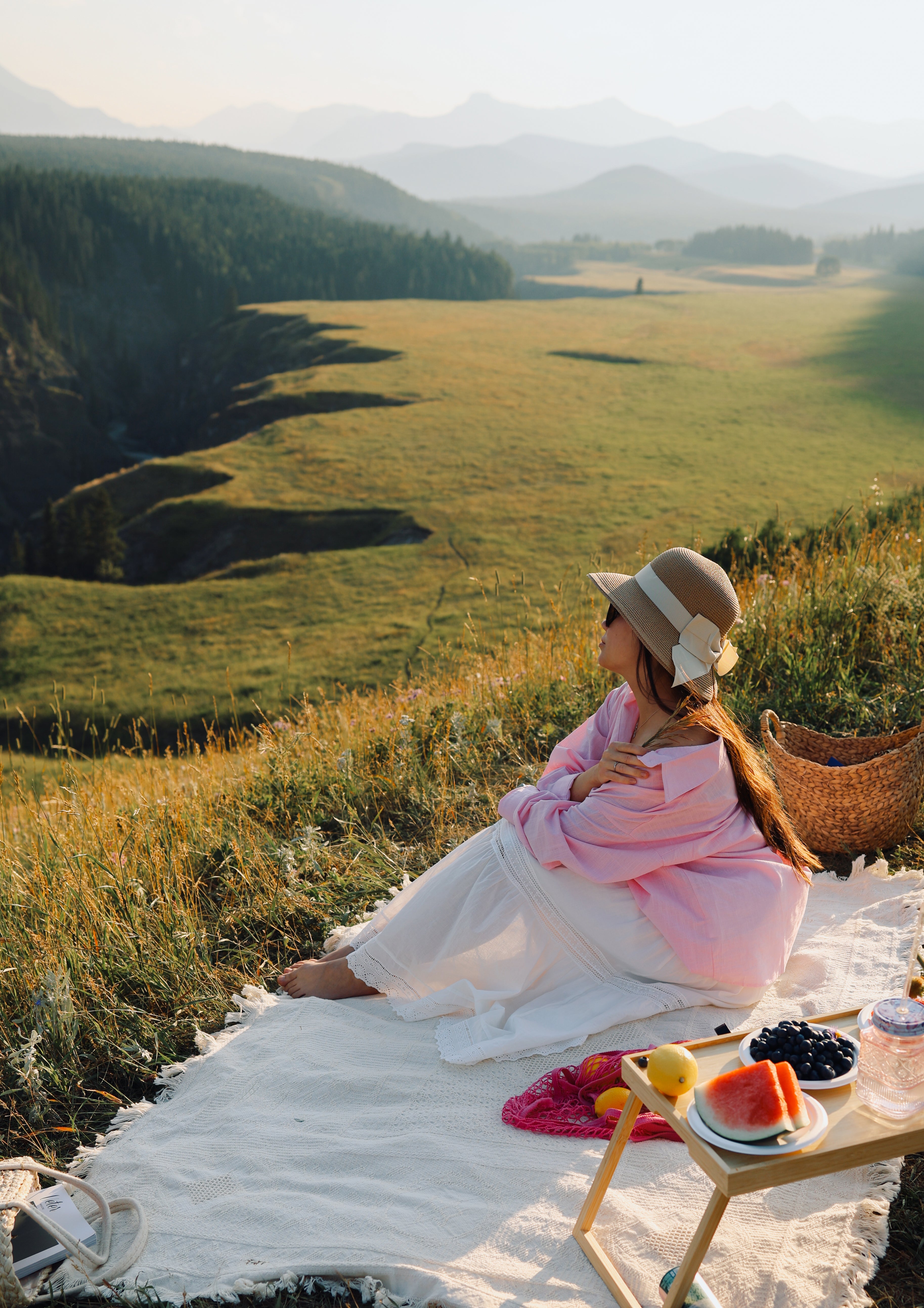 A woman in a pink kimono and straw hat sits on a white blanket in a mountain meadow, with a small picnic table holding fruit and a basket nearby.