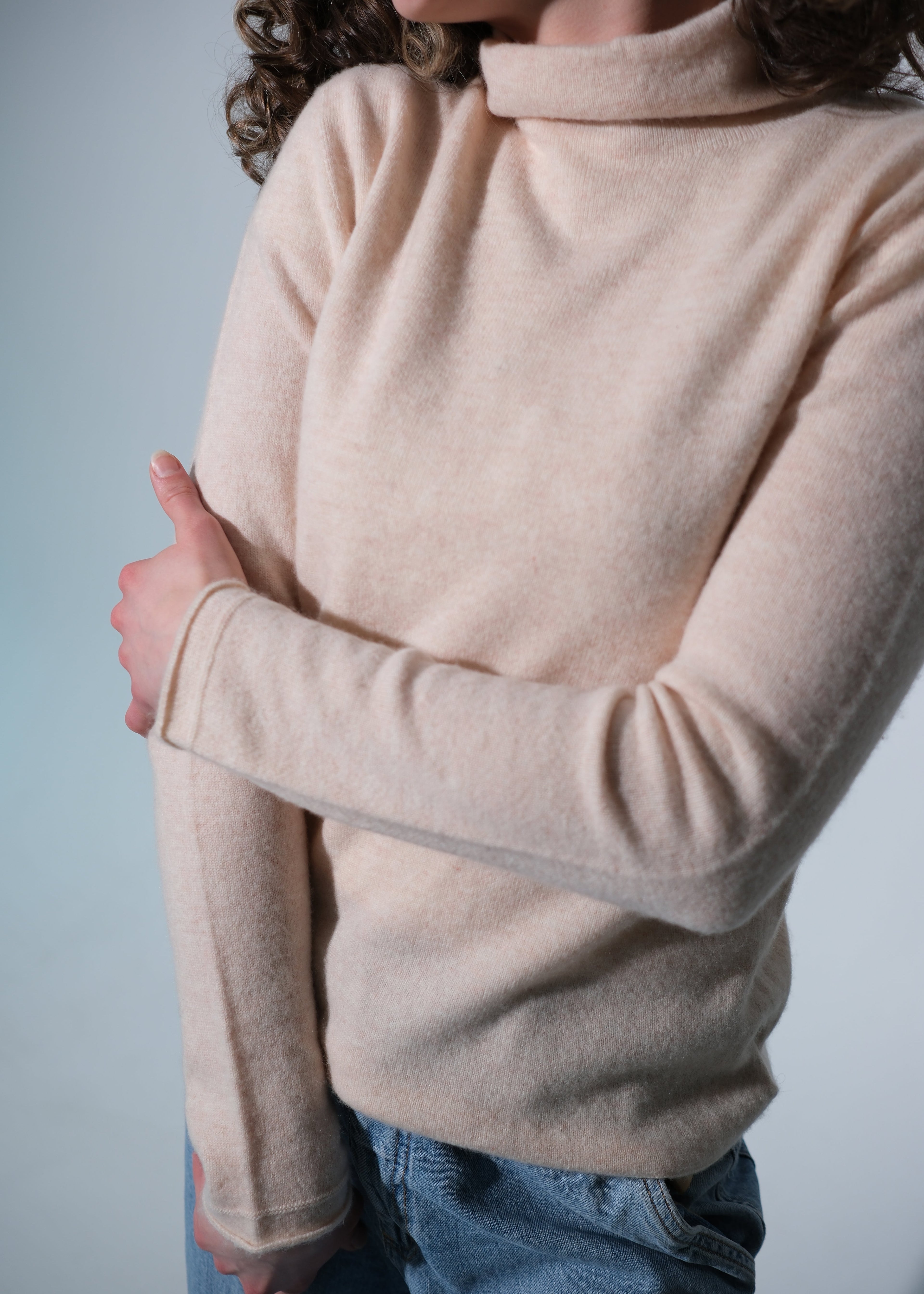 A close-up of a person wearing a light beige, long-sleeve mock neck sweater with blue jeans, set against a plain light gray background.