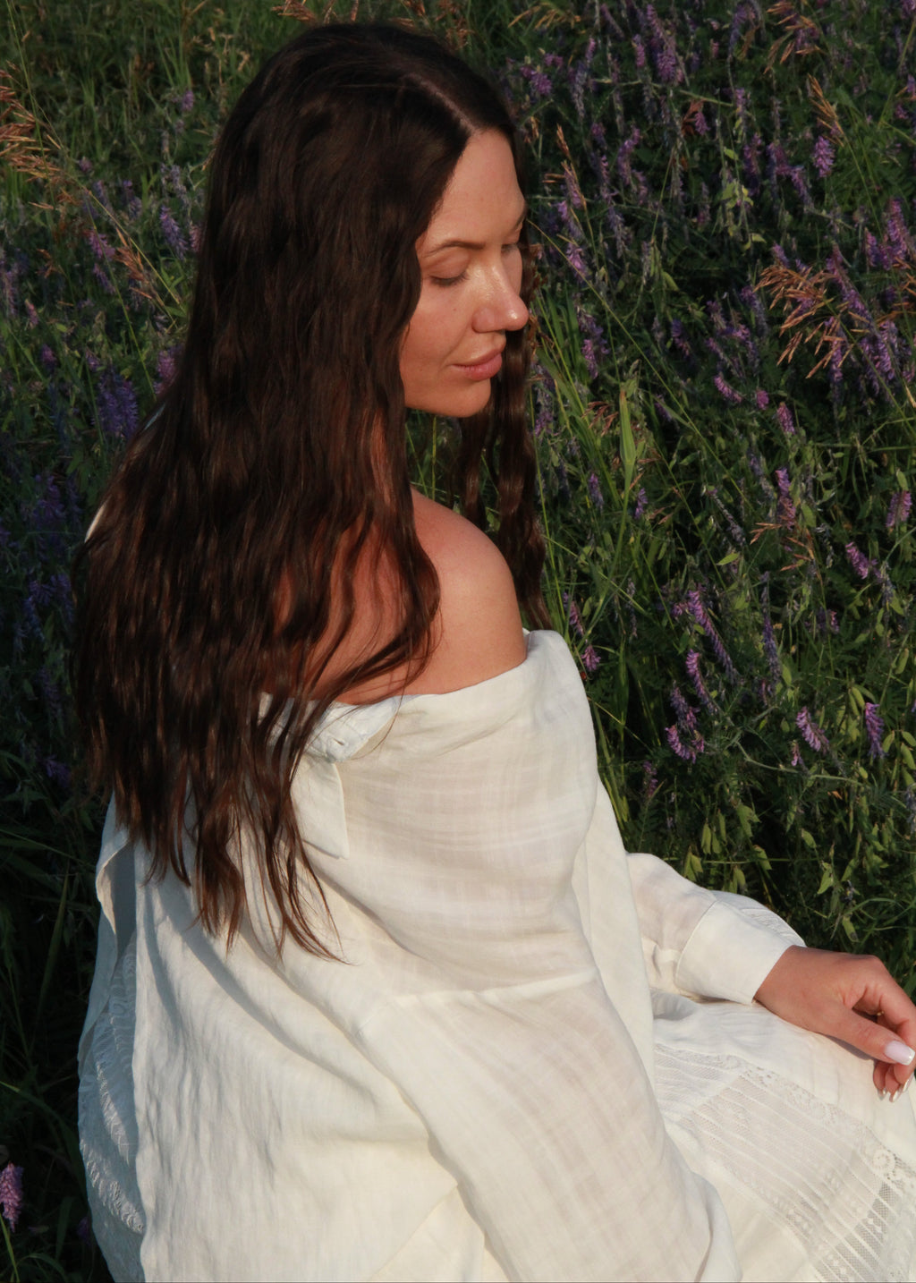 A woman with long dark brown hair sits outdoors in a field of purple flowers, wearing a loose off-the-shoulder white linen dress or shawl.