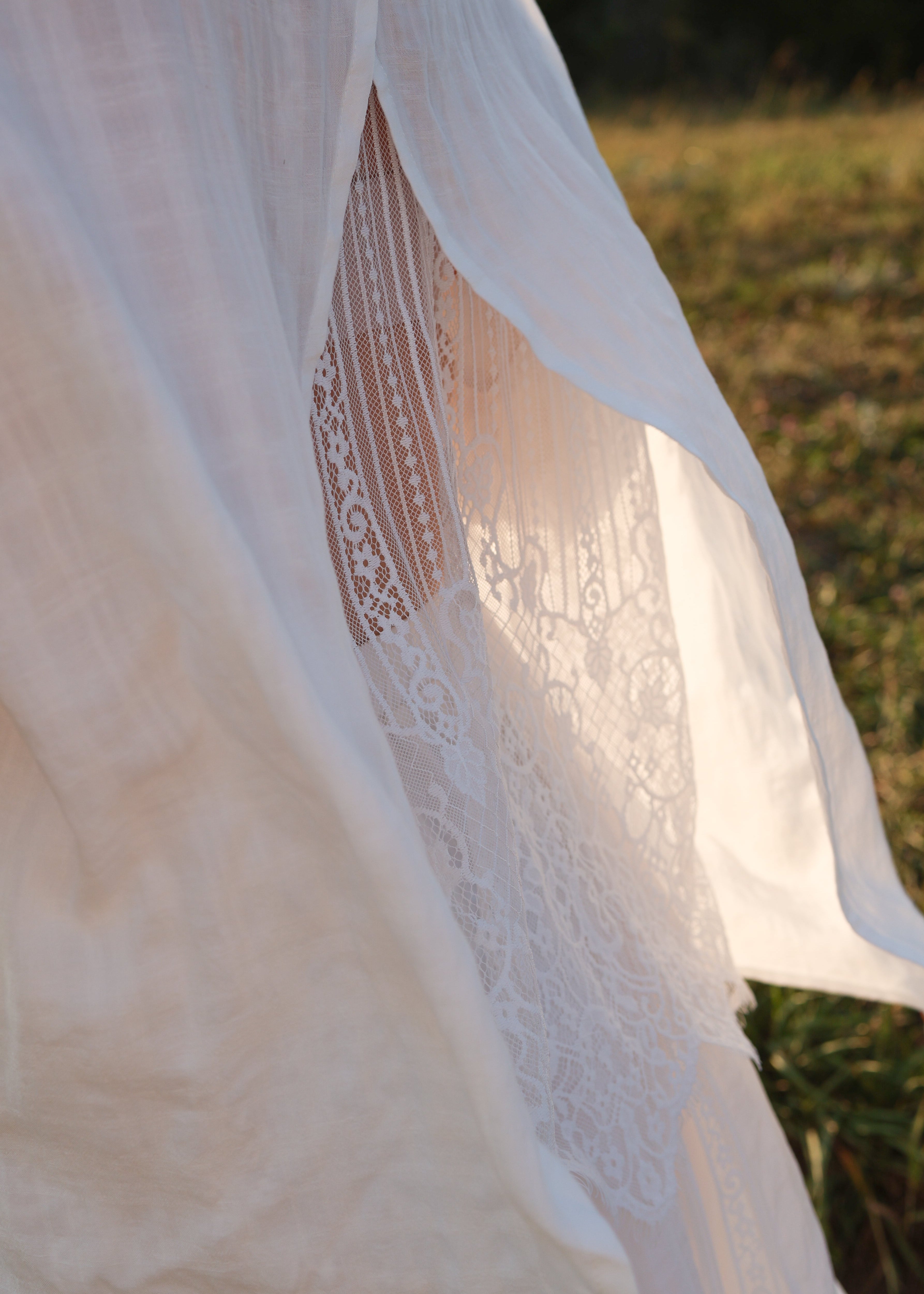 A close-up of a flowing white sheer fabric garment or drape with delicate lace detailing, photographed outdoors against a softly blurred grassy background.
