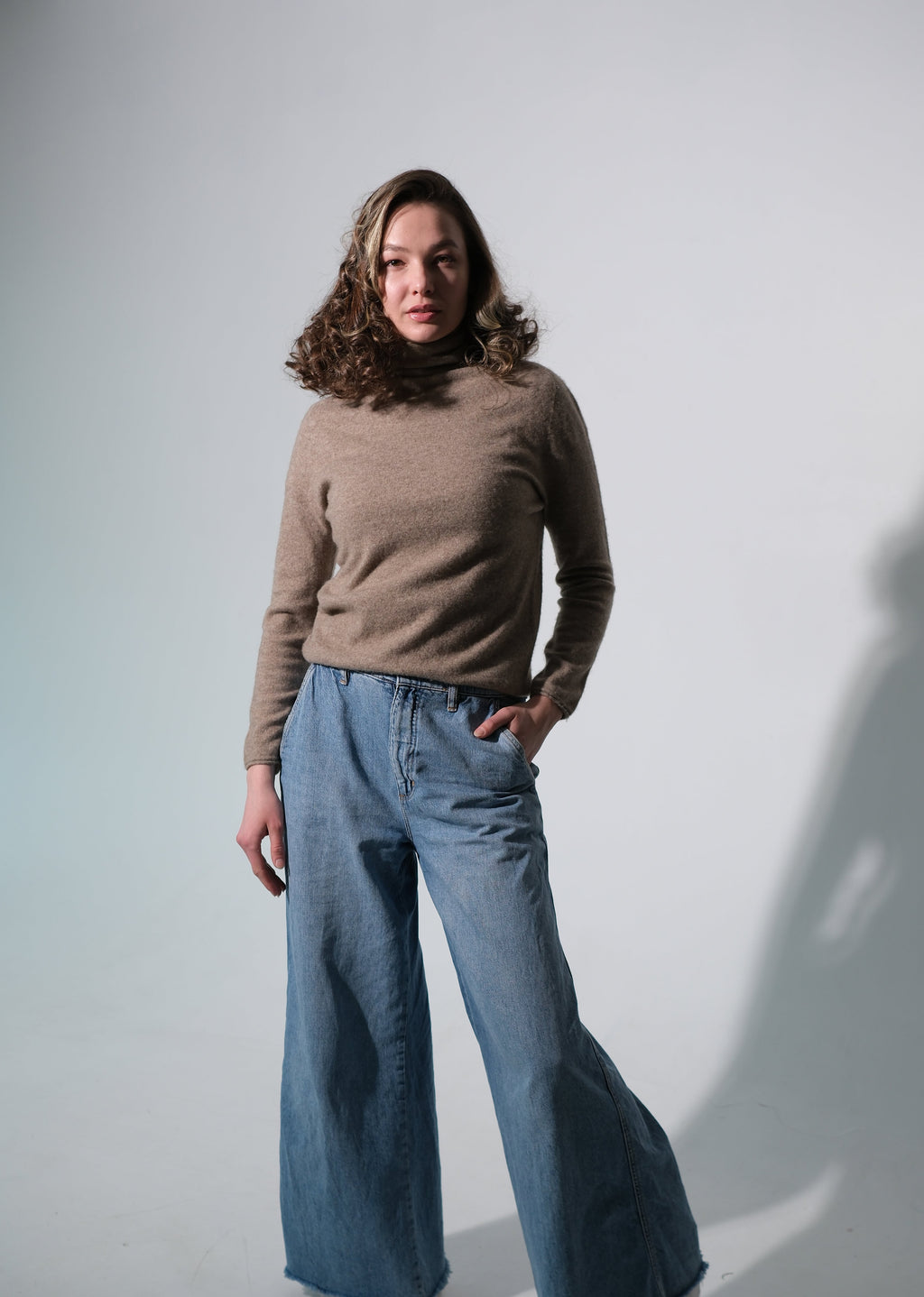 A woman modeling a beige mock-neck knit sweater and wide-leg blue jeans against a plain light gray studio background with a strong cast shadow.
