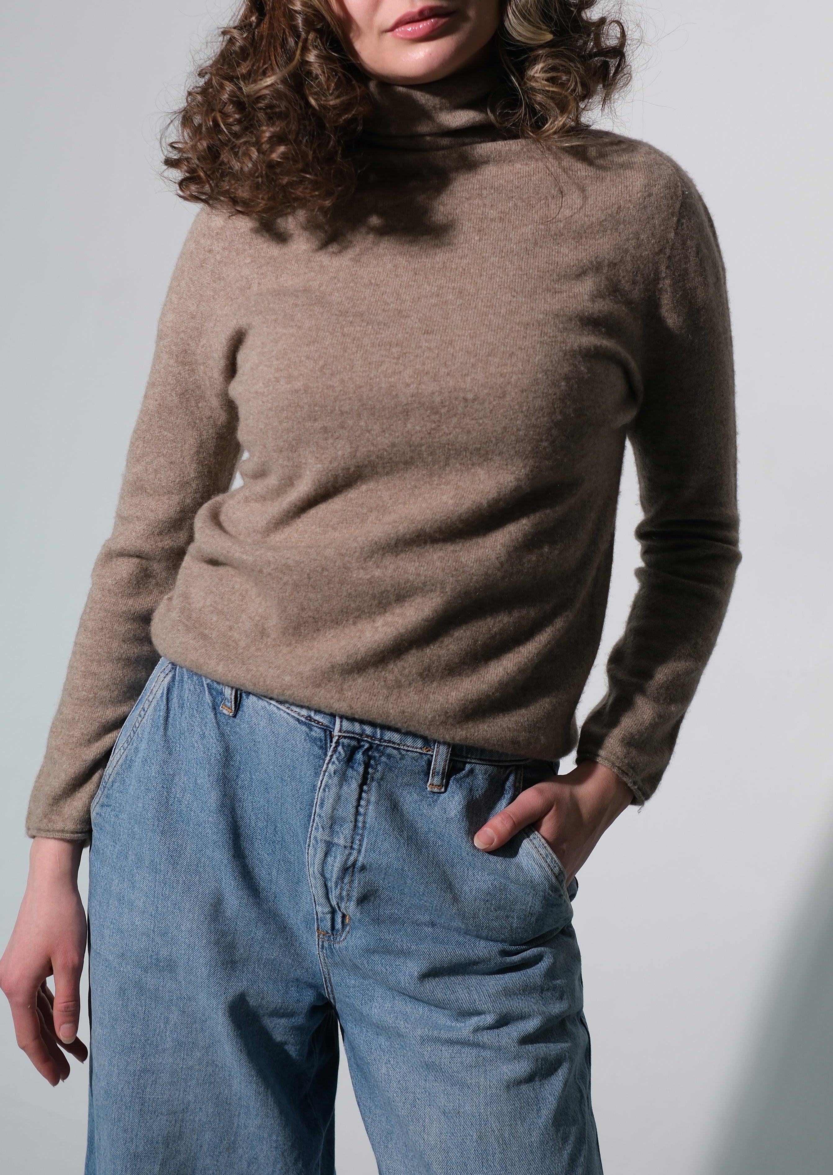 A woman with wavy brown hair wearing a beige turtleneck sweater and wide-leg blue jeans, standing against a light gray studio background with a soft shadow cast to the right.