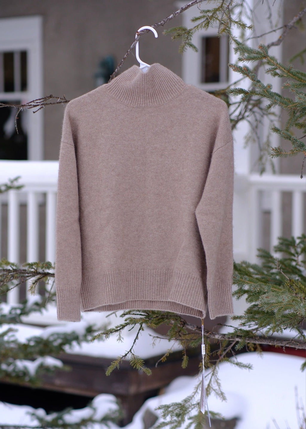 A light taupe knit mock neck sweater hanging from a white plastic hanger on a tree branch in a snowy outdoor setting.