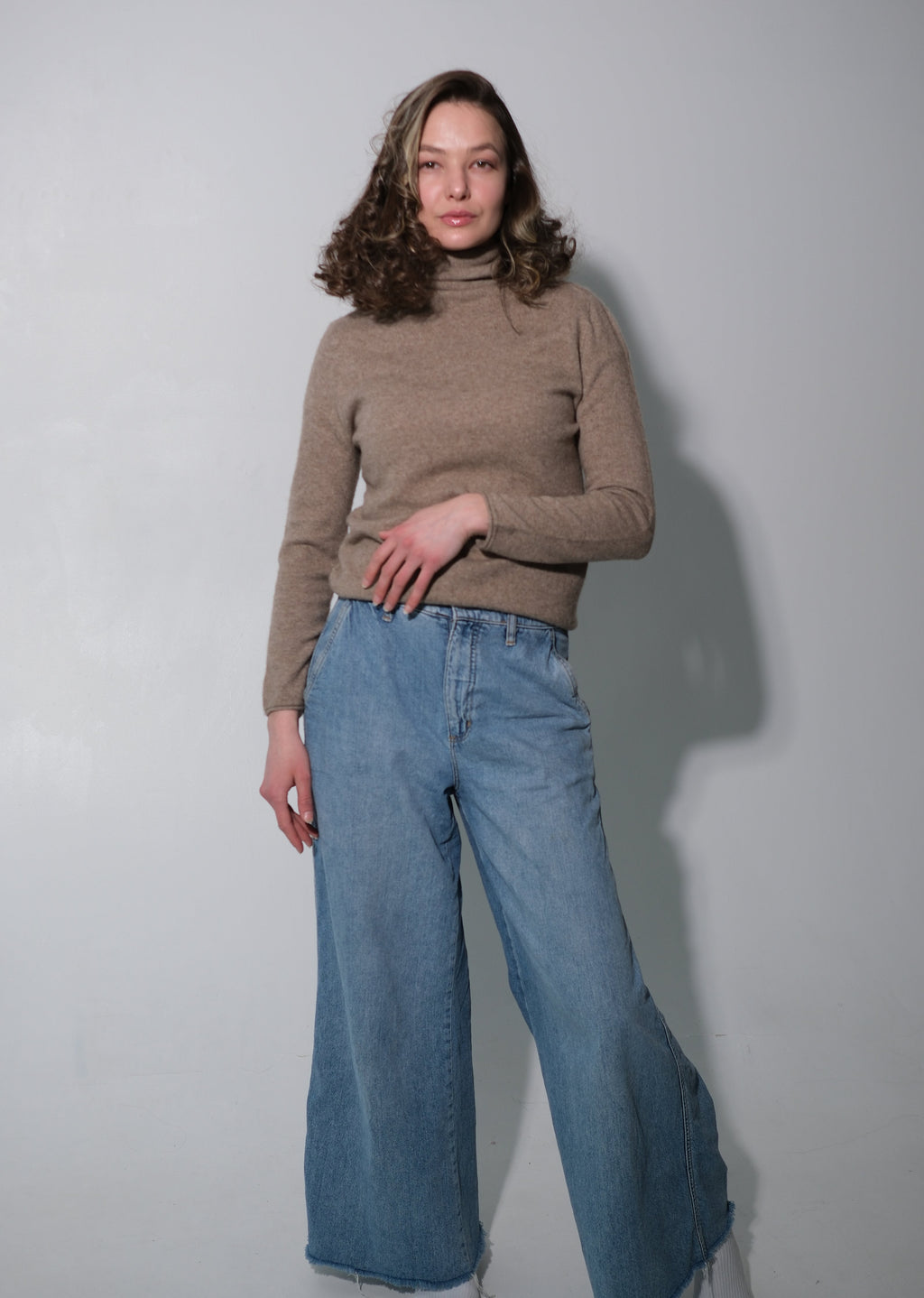 A woman modeling a beige ribbed turtleneck sweater and wide-leg blue denim jeans against a plain light gray studio background.