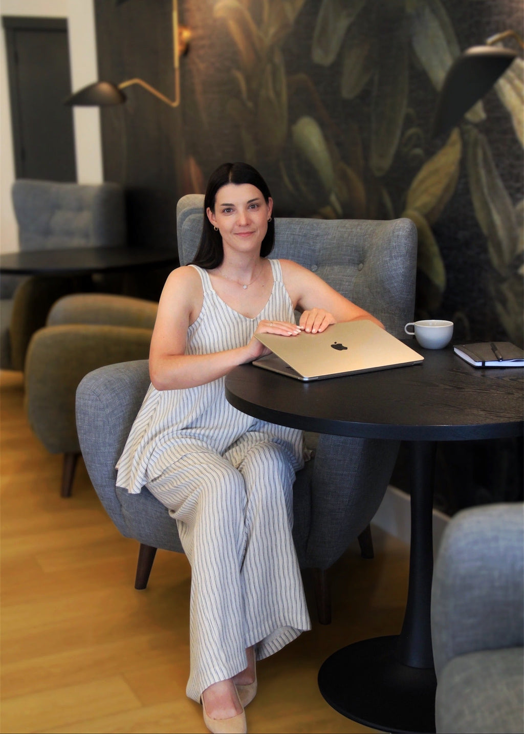 A woman sits in a gray armchair at a round black table, wearing a light blue and white vertically striped sleeveless jumpsuit. She has a silver laptop on the table in front of her, with a white mug and a smartphone nearby.