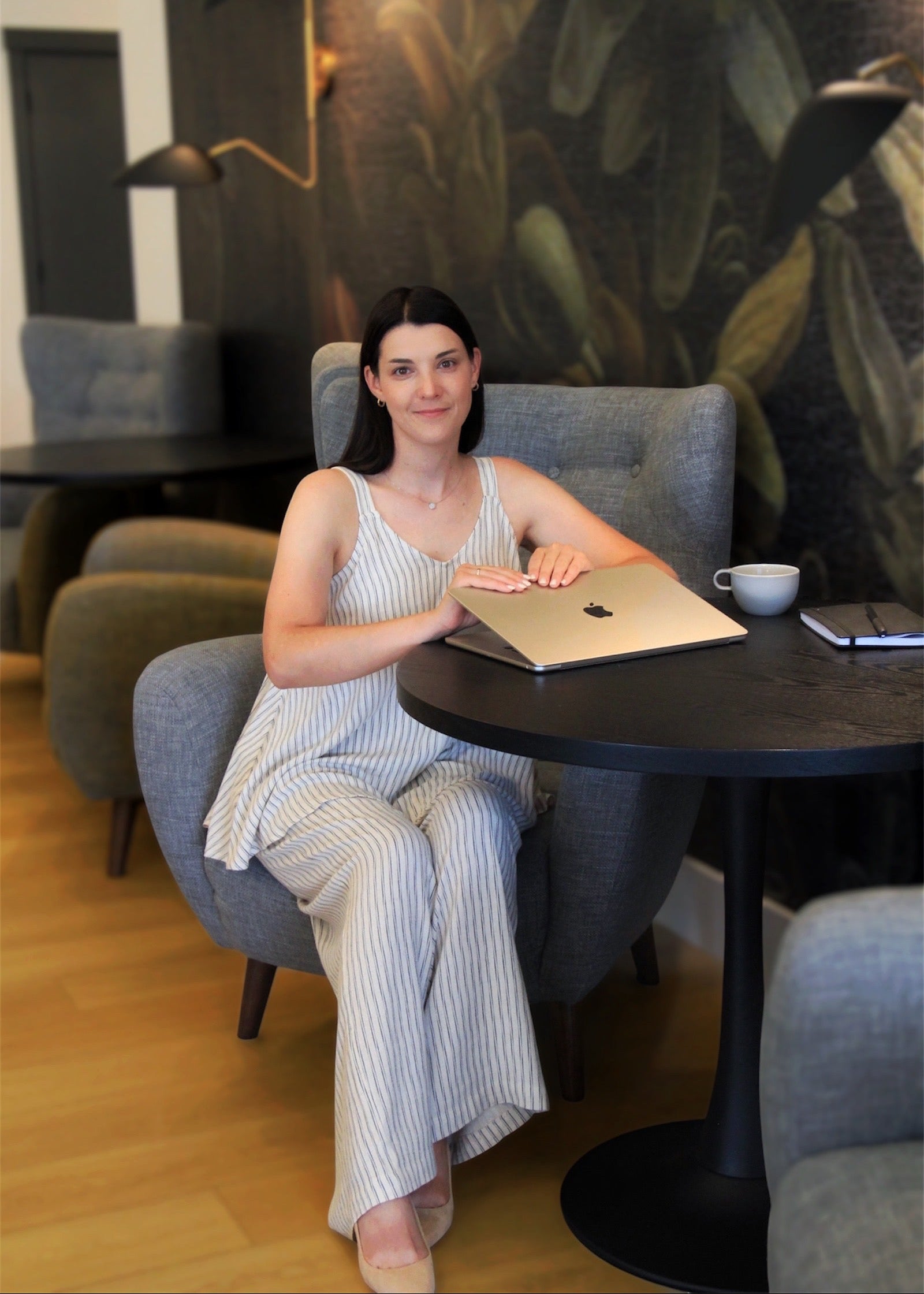 A woman sits in a gray armchair at a round black table, wearing a light blue and white vertically striped sleeveless jumpsuit. She has a silver laptop on the table in front of her, with a white mug and a smartphone nearby.