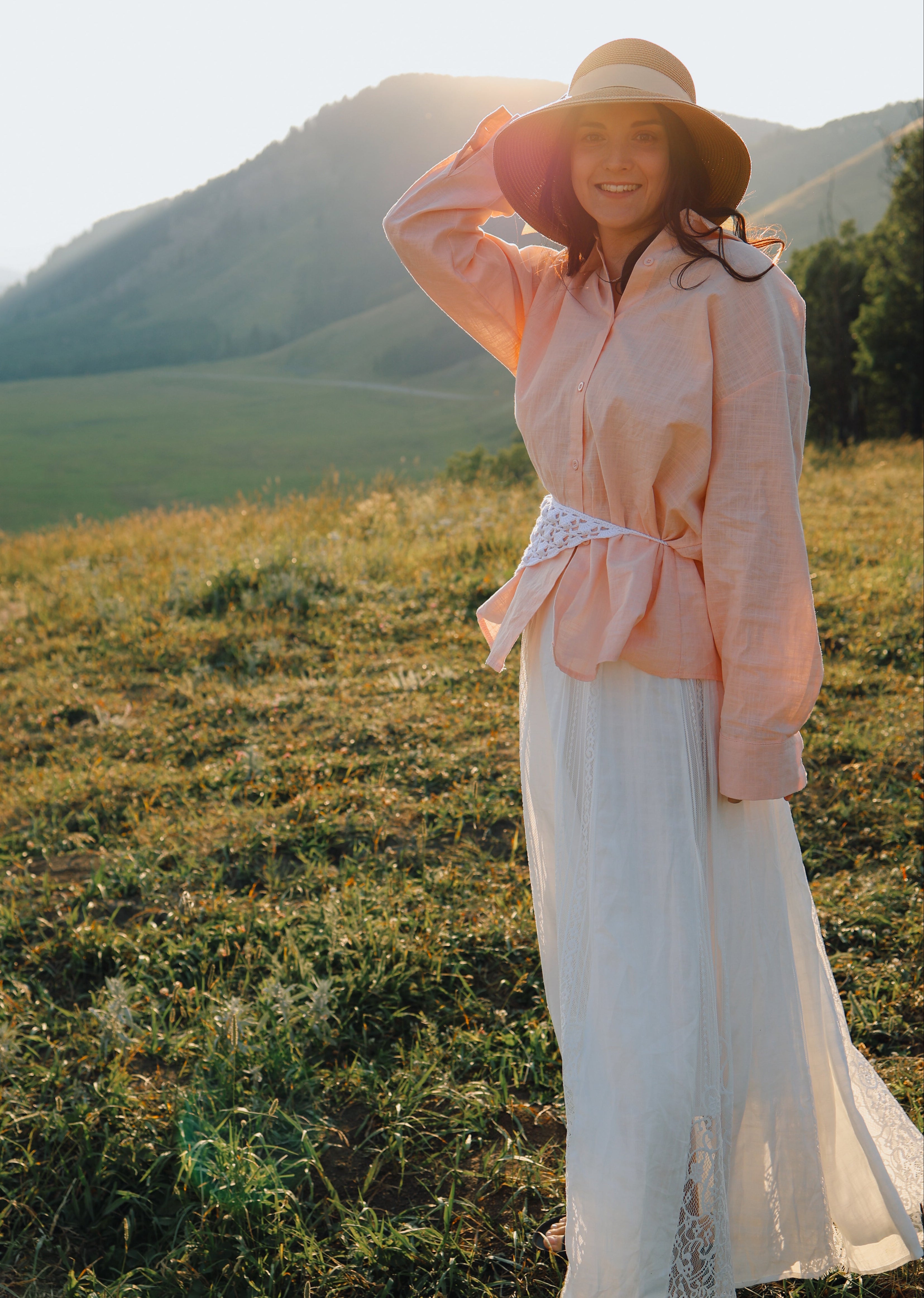 A woman stands outdoors in a grassy field with rolling hills in the background, wearing a light pink long-sleeve blouse tied at the waist, a long white skirt, and a wide-brimmed straw hat.
