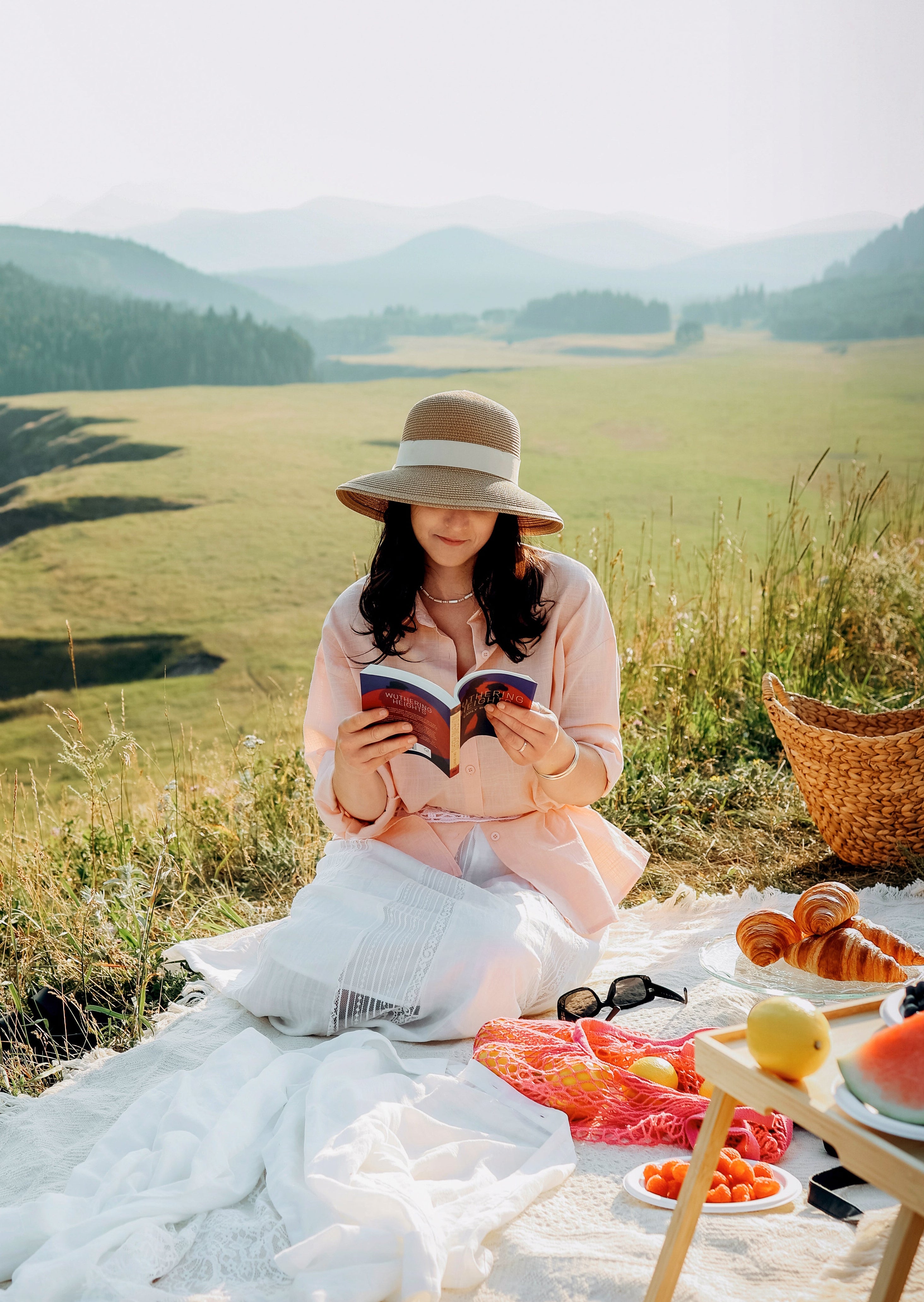 A woman in a pink dress and wide-brimmed hat sits on a white blanket in a grassy mountain meadow, reading a book. A picnic setup with a basket, bread, fruit, and sunglasses is arranged beside her.