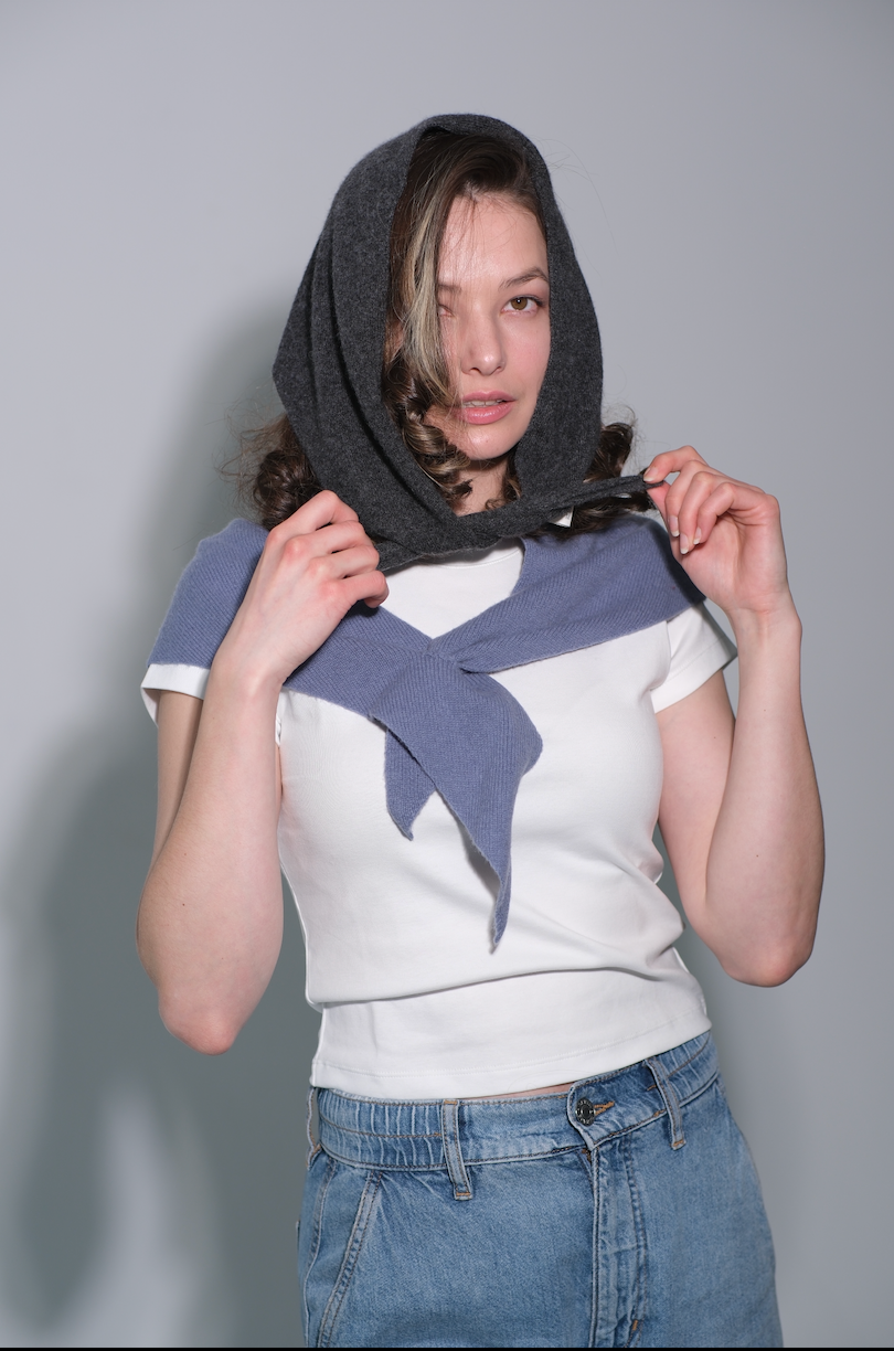 A woman wearing a gray knit headscarf tied around her neck over a white t-shirt and blue jeans, posing against a solid gray studio background.