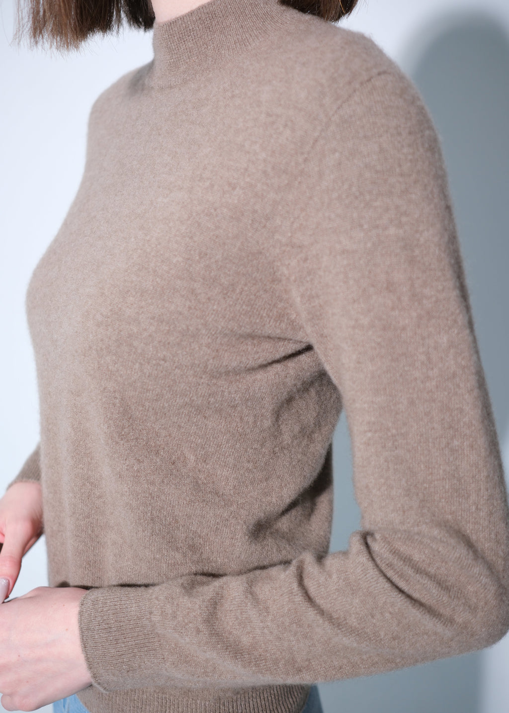 A close-up of a person wearing a taupe brown knit mock neck sweater, showing the texture and fit against a light gray background.