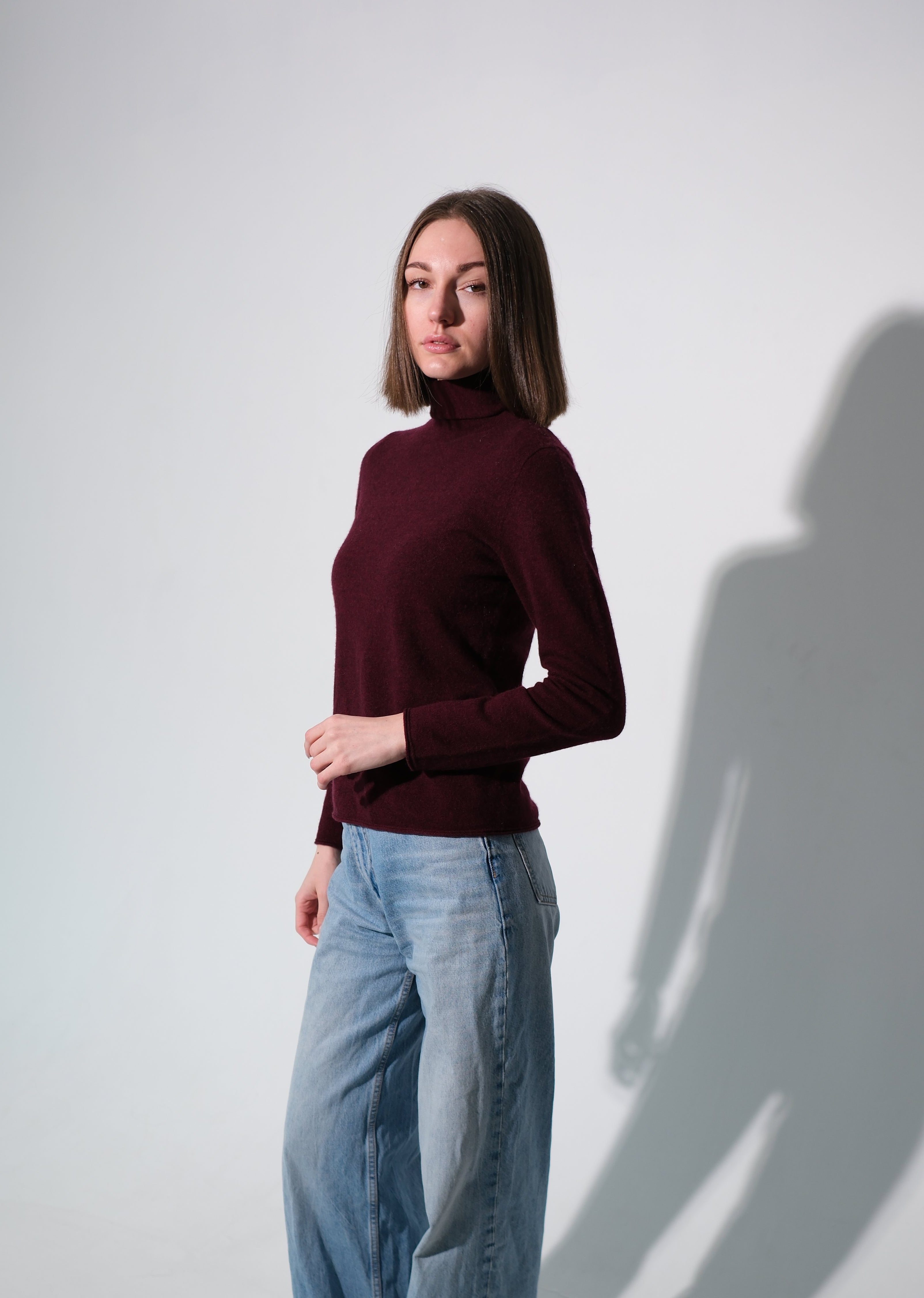 A woman modeling a dark burgundy mock neck knit sweater paired with wide-leg light blue jeans, standing against a light gray studio wall with a prominent shadow.