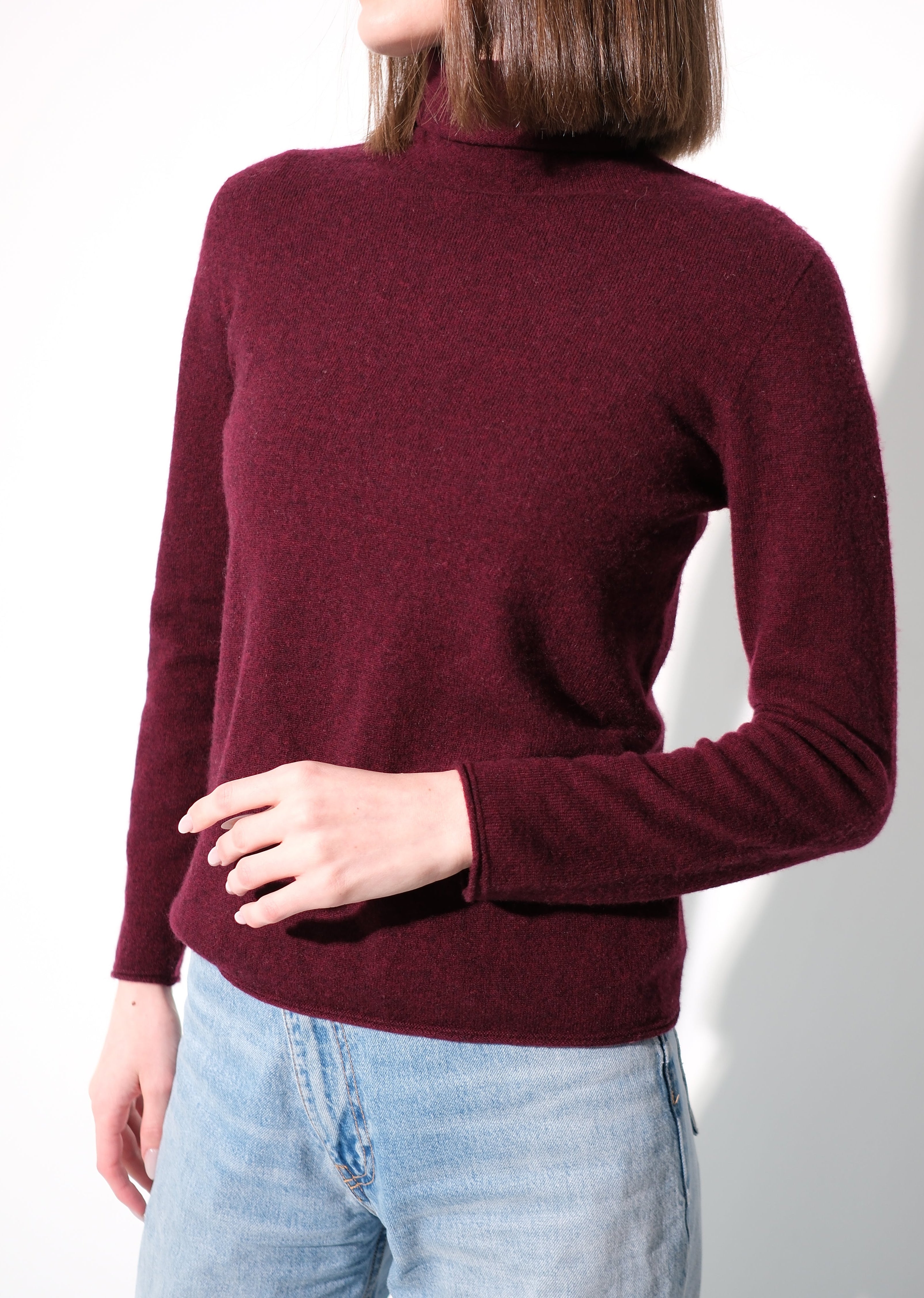 A woman wearing a long-sleeve ribbed knit mock neck sweater in burgundy, paired with light wash blue jeans, standing against a plain white wall.