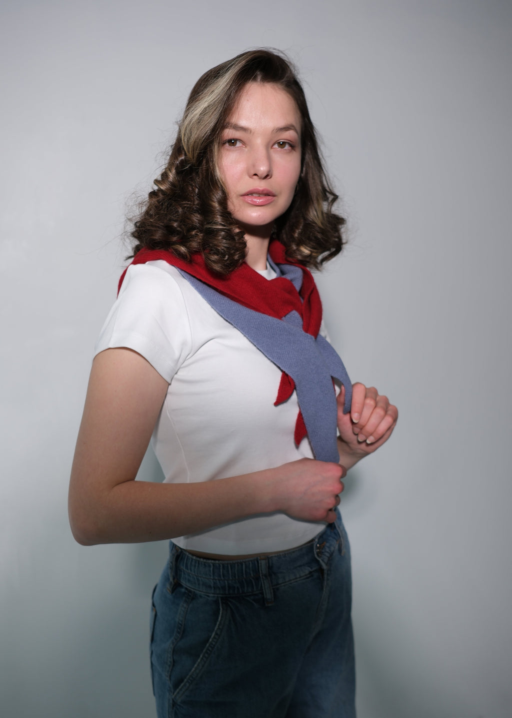A woman modeling a white short-sleeve knit top with a red and gray geometric pattern, paired with blue jeans, against a plain gray studio background.