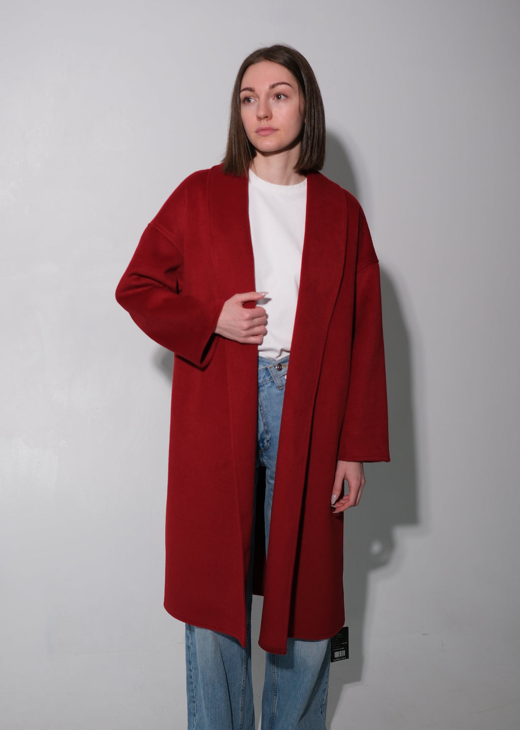 A woman stands against a plain light gray wall wearing a long, open-front red coat over a white top and light blue jeans.