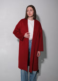 A woman stands against a plain light gray wall wearing a long, open-front red coat over a white top and light blue jeans.