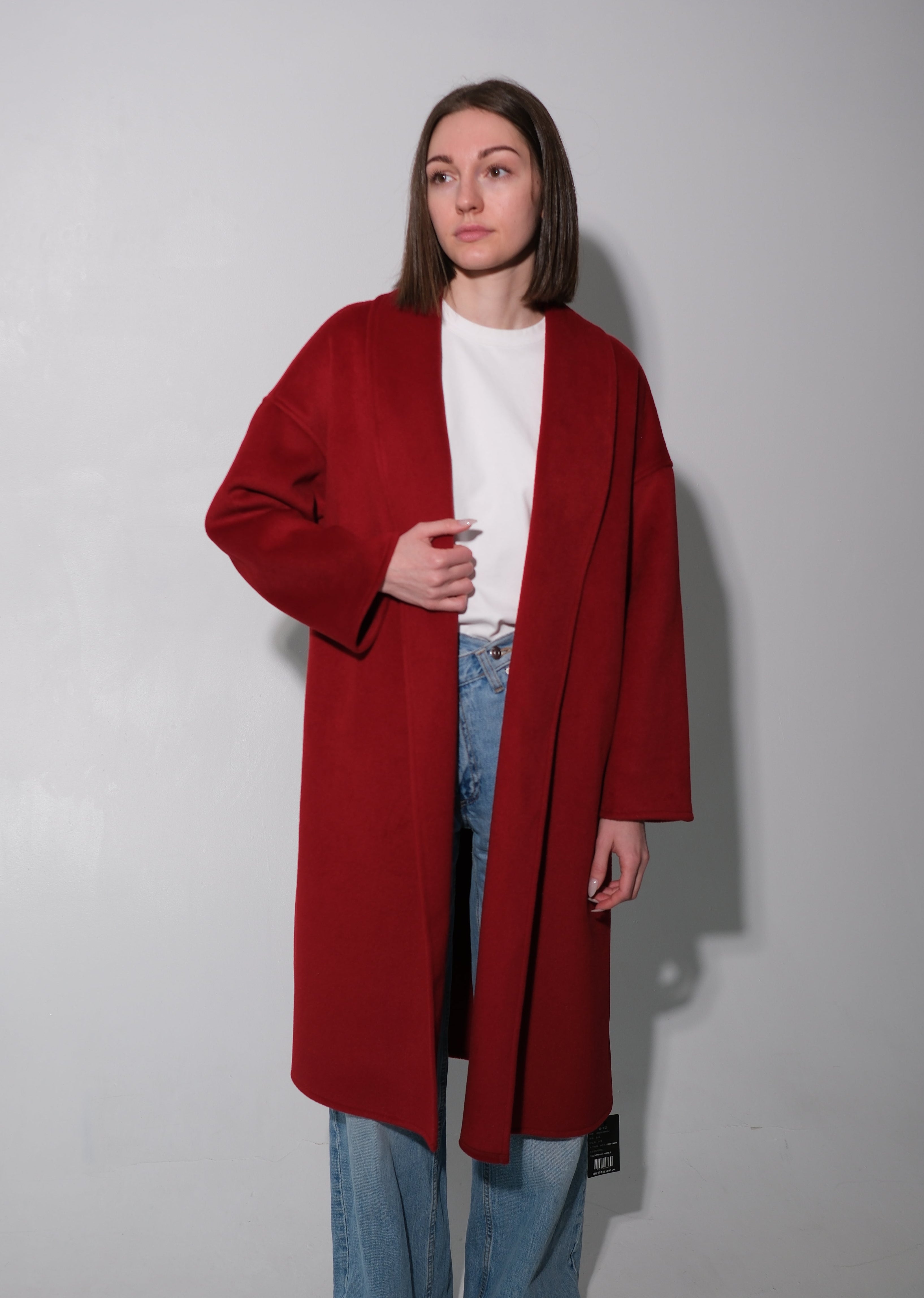 A woman stands against a plain light gray wall wearing a long, open-front red coat over a white top and light blue jeans.