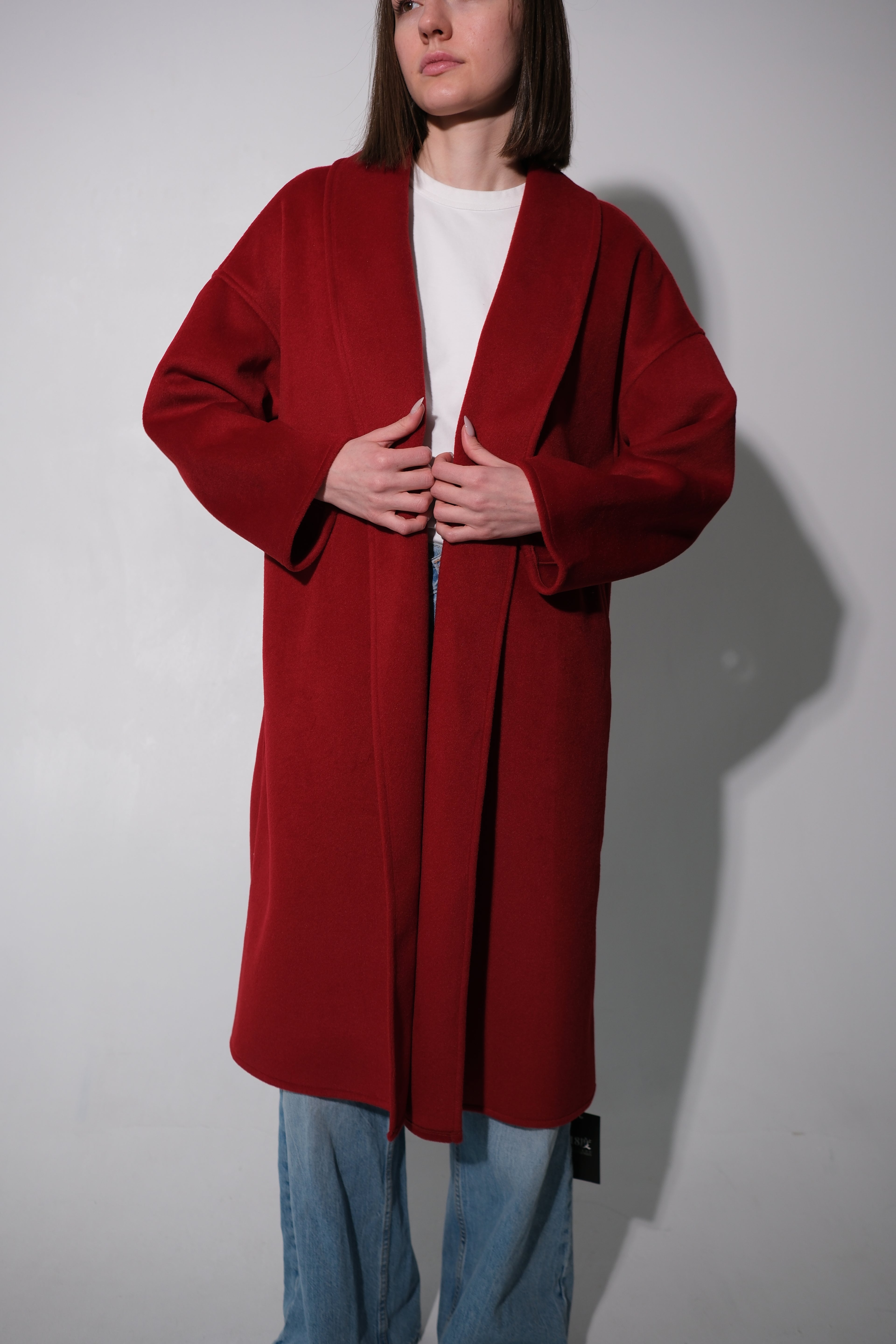 A woman wearing a long, open-front red coat over a white top and blue jeans, standing against a plain light gray wall.