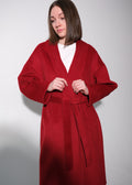 A woman wearing a long, wide-sleeved red wrap coat with a matching tie belt over a white top, standing against a plain light gray studio background.