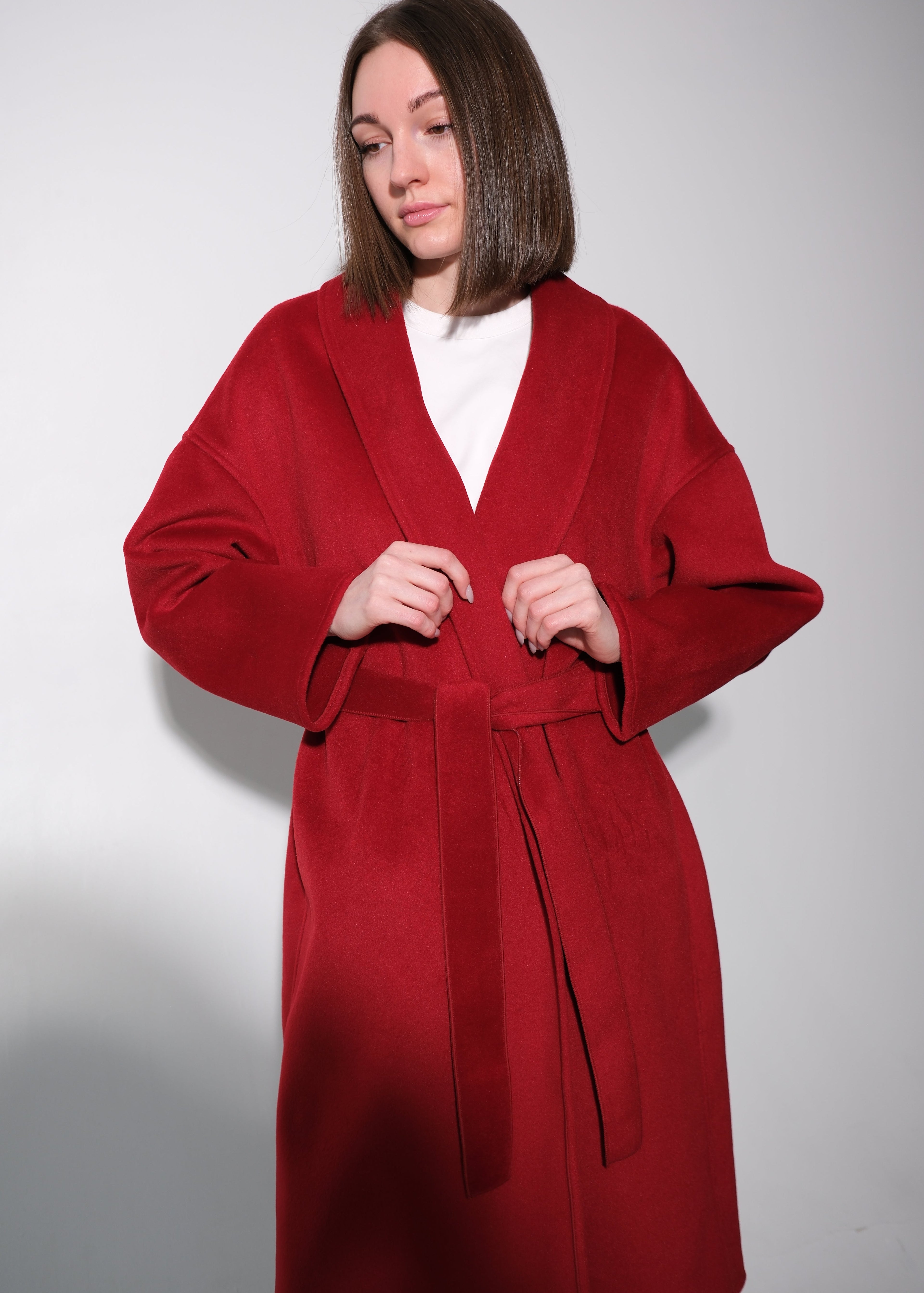 A woman wearing a long, wide-sleeved red wrap coat with a matching tie belt over a white top, standing against a plain light gray studio background.